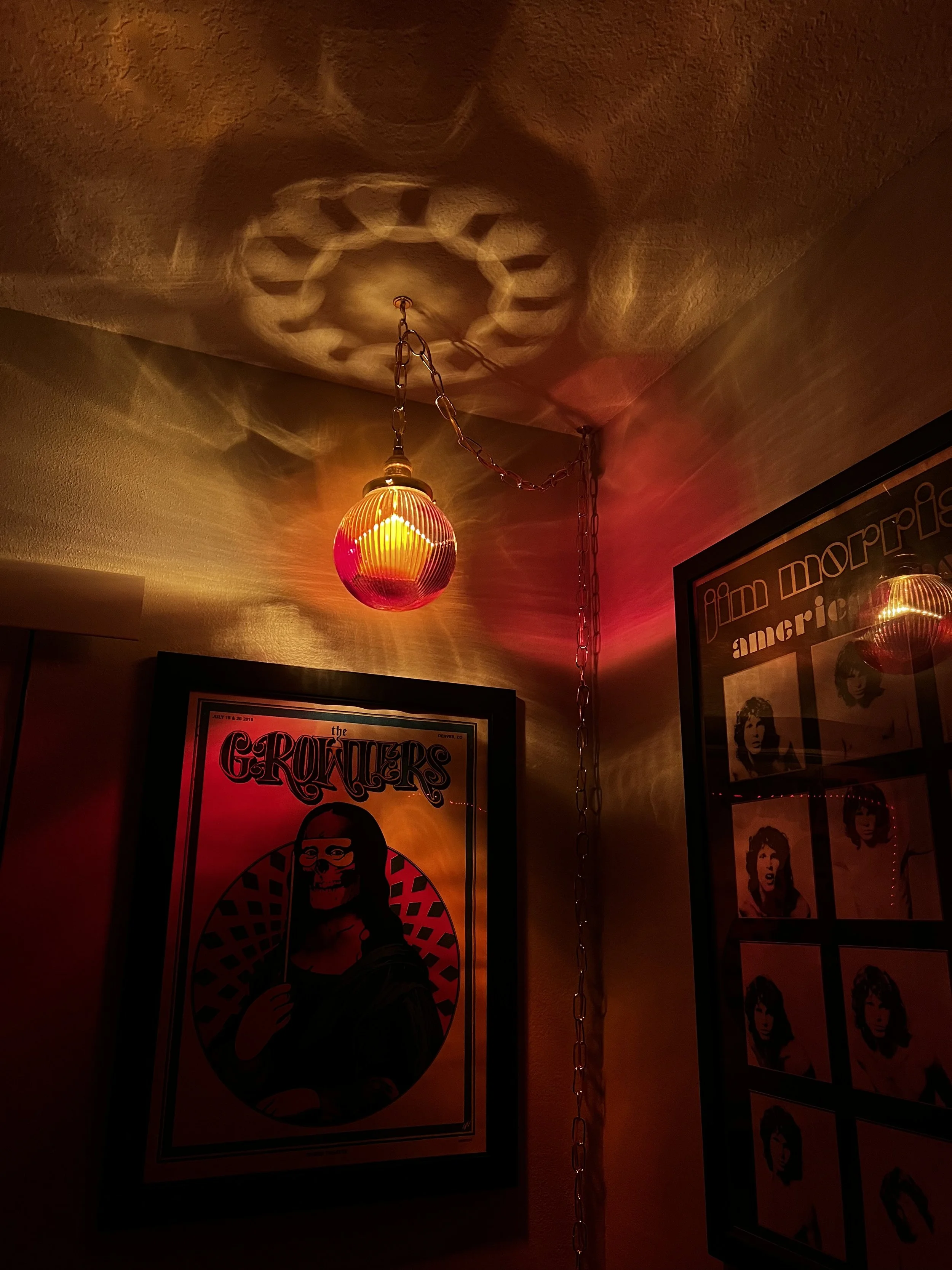 Room with a red and yellow hanging resin lamp casting patterned shadows on the ceiling, and framed posters of The Growlers and Jim Morrison on the wall.
