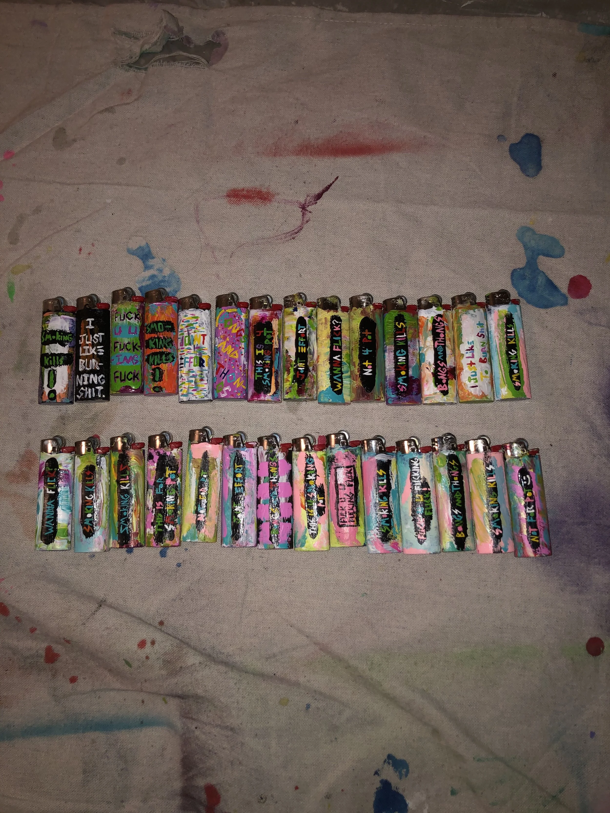Two rows of colorful, decorated lighters on a paint-splattered cloth background.