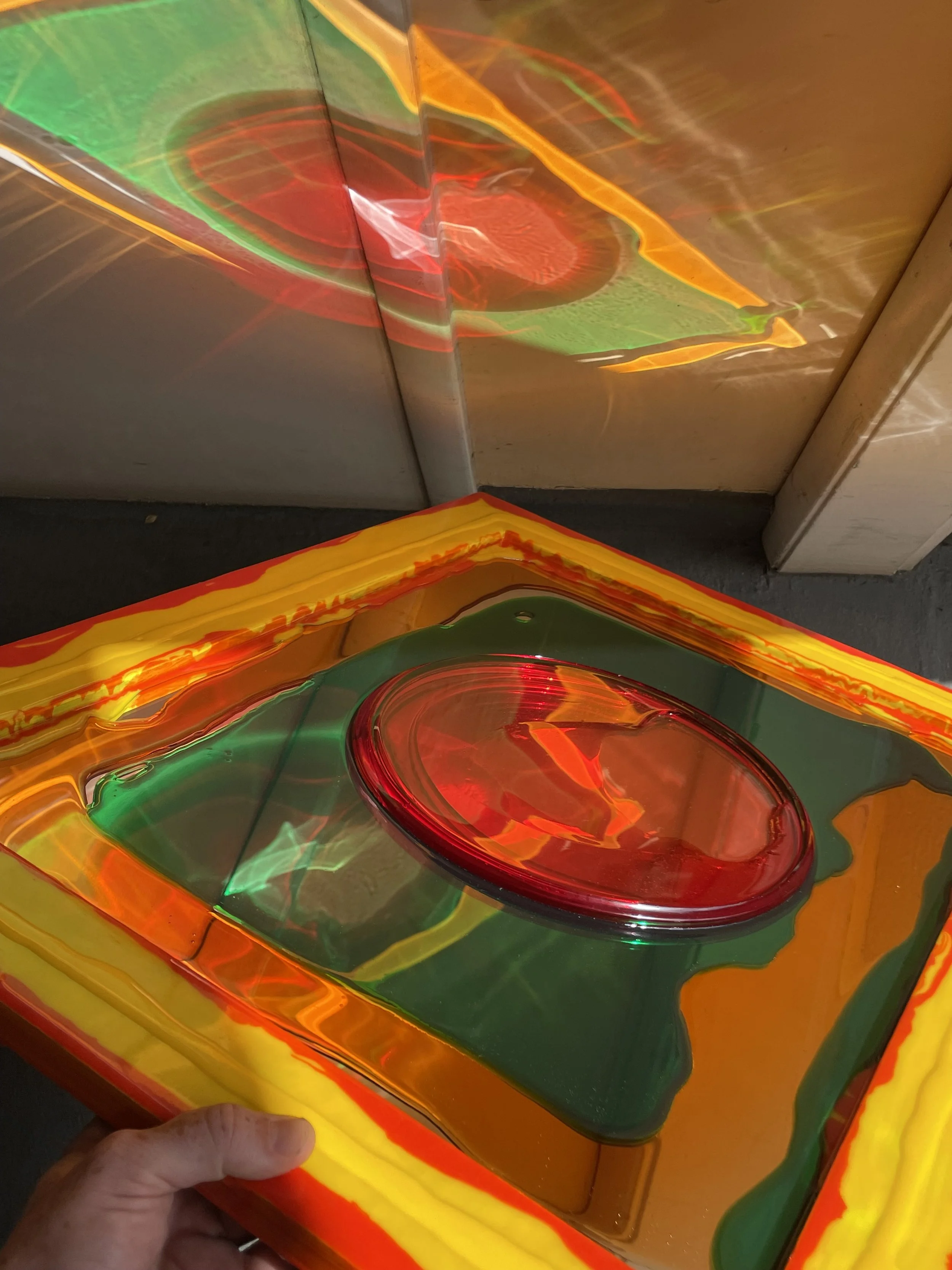 Multicolored resin mirror  with a red circle shape in the middle, reflecting light, placed inside a vibrant frame of yellow, red, green, and orange layers.