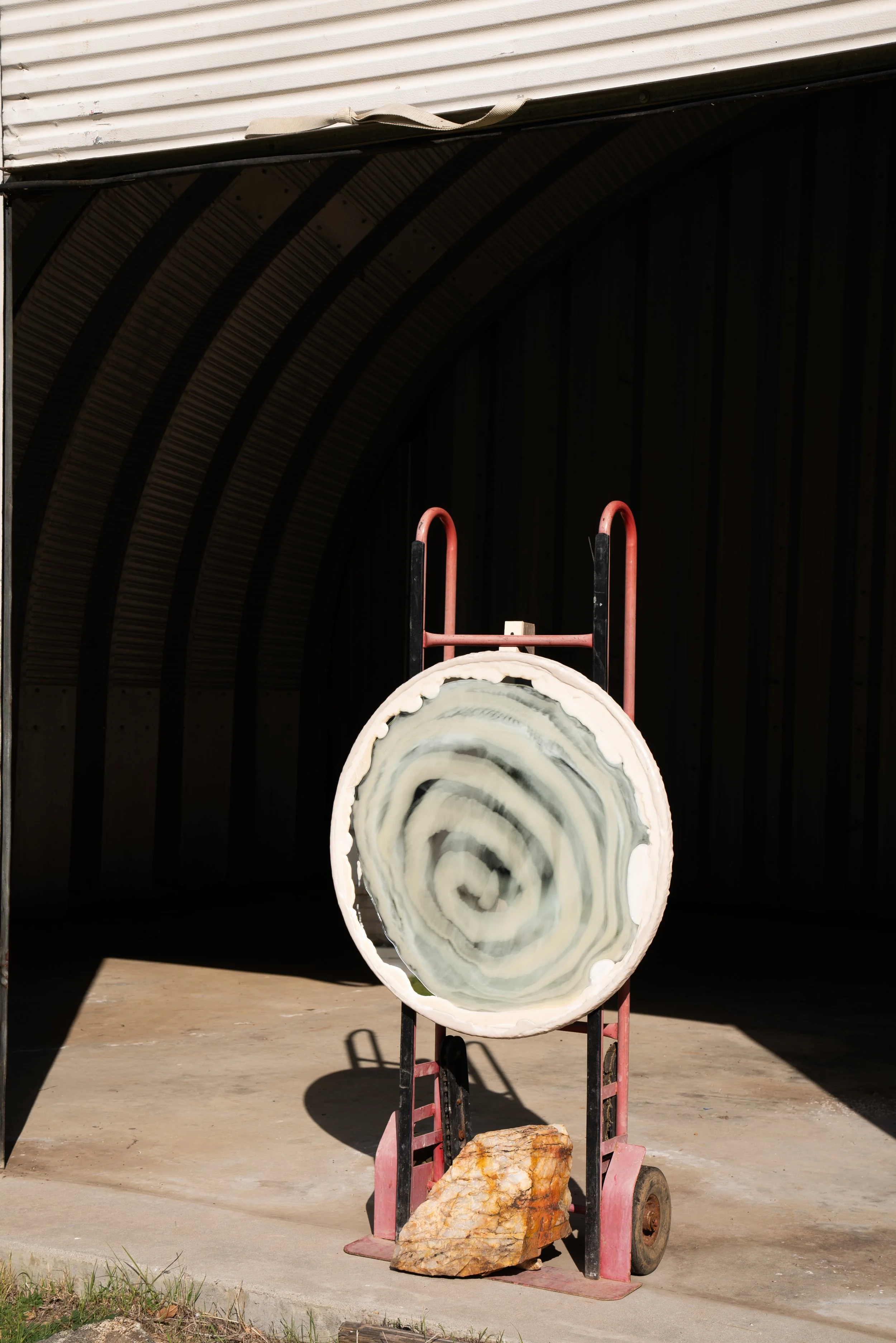 A large circular resin mirror with a swirl pattern, mounted on a dolly outside a metal building.