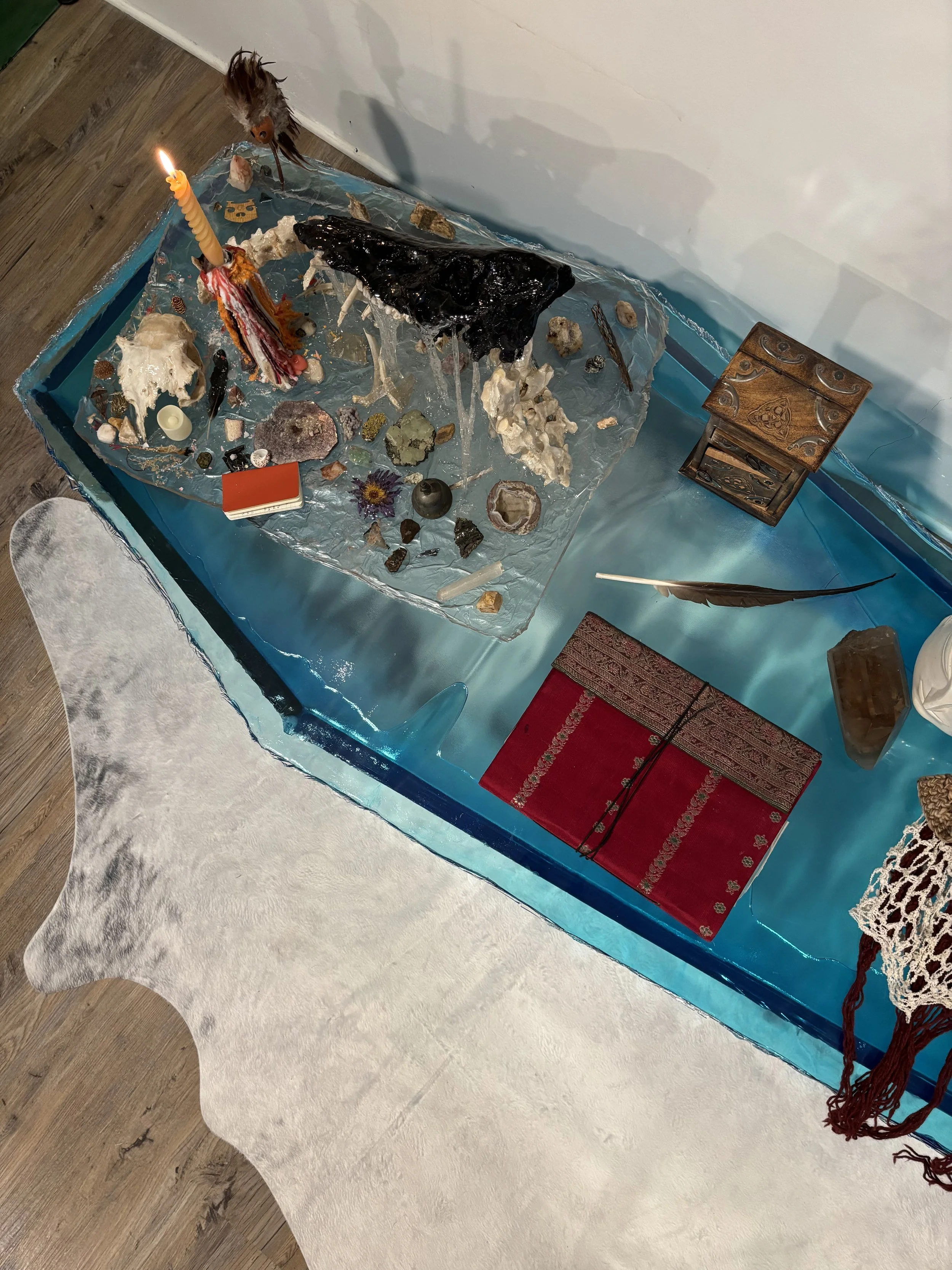 A resin altar with various objects including a lit candle, rocks, bones, feathers, a red book, and decorative items, set on a blue resin coffee table with a white furry rug nearby.