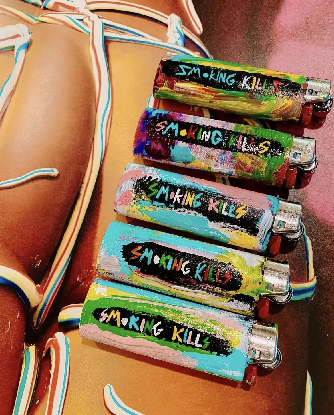 Colorfully painted lighters with the words 'SMOKING KILLS' written on them in various colors, placed on a surface beside a decorated object.