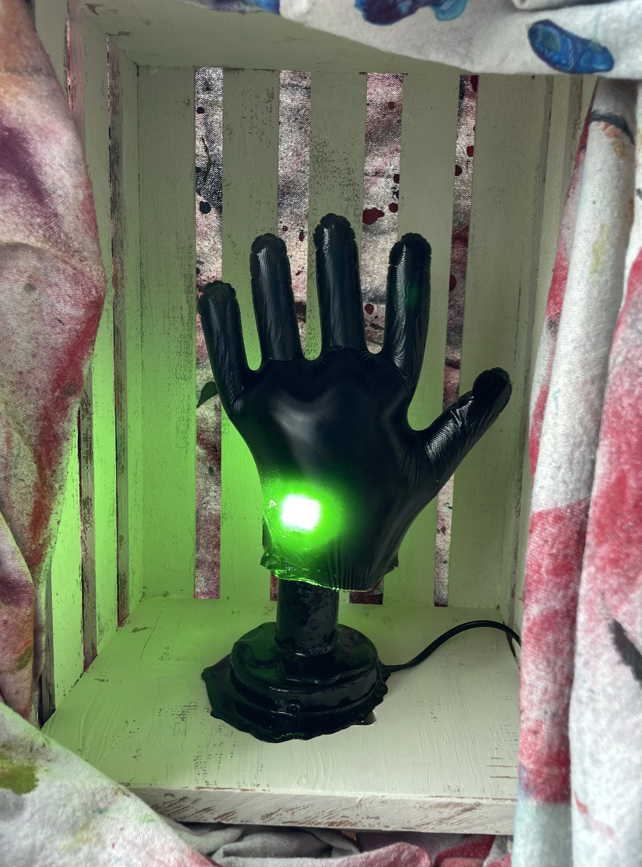 A black hand-shaped resin lamp with a green light glowing from the palm is placed inside a wooden box with slats. The box is surrounded by curtains with paint splatters.