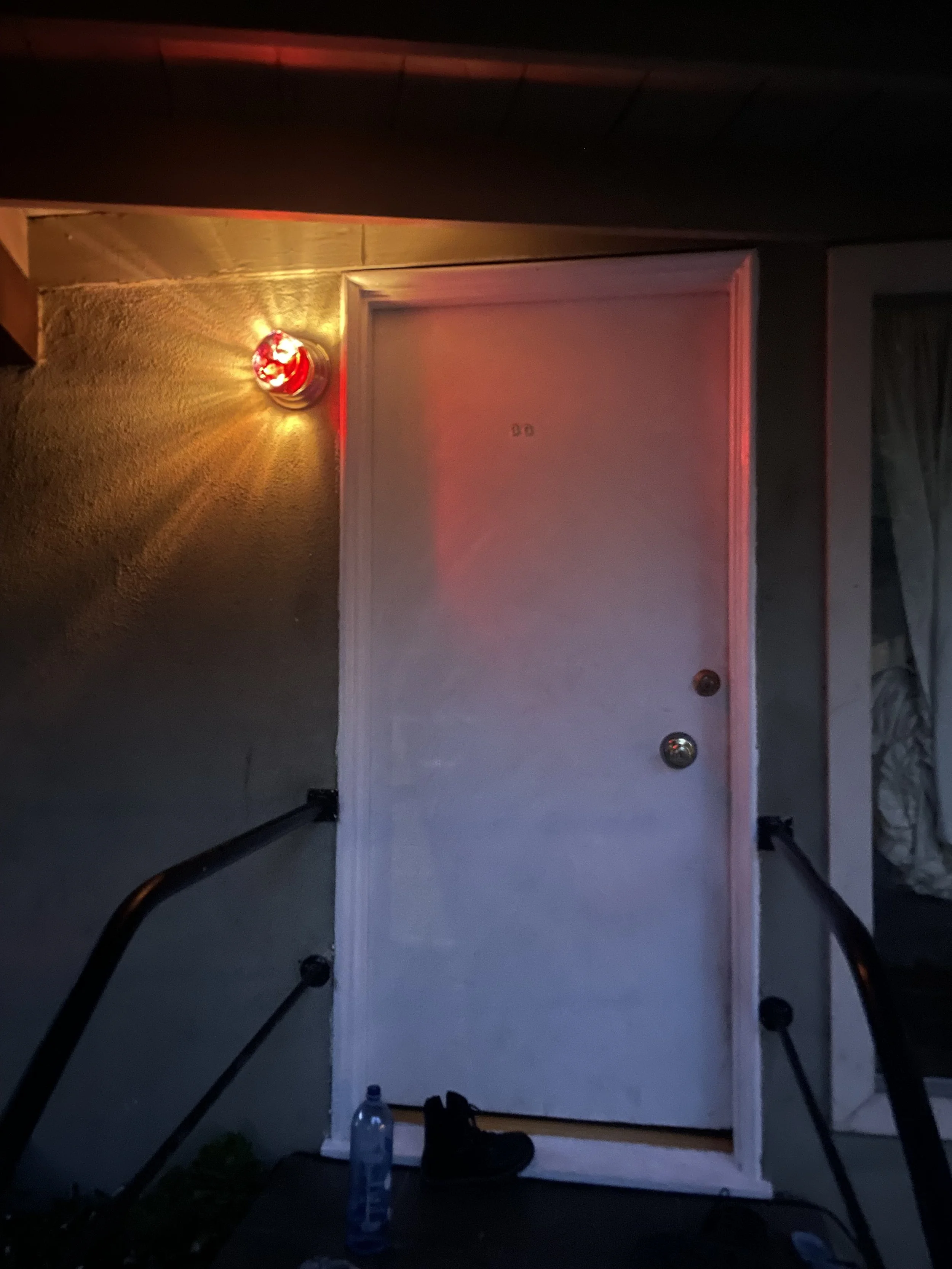 Front door of a house with a red resin light above it, a black shoe, and a water bottle on the ground outside.