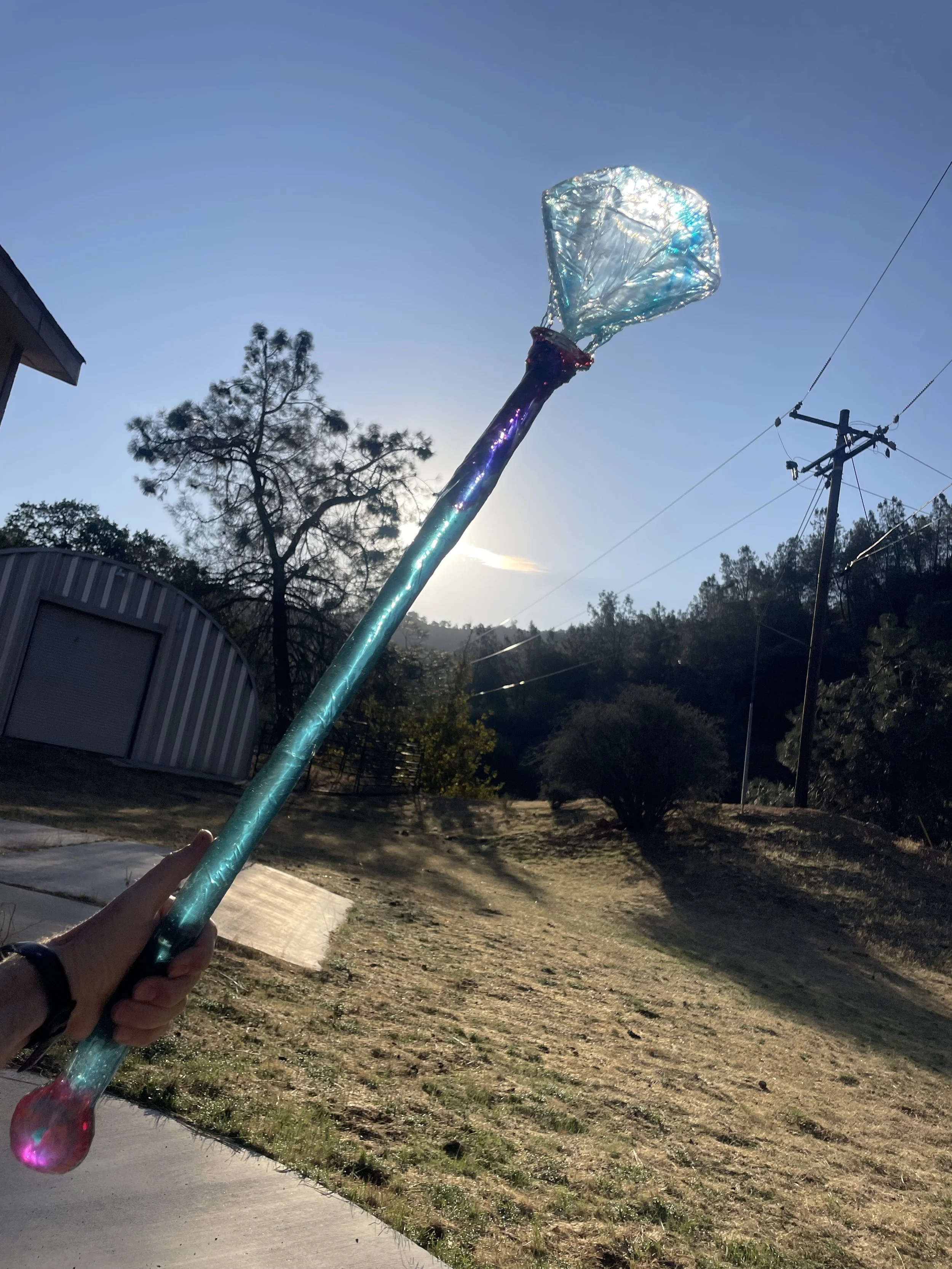 Person holding a large, colorful, resin magic wand or scepter with a transparent, iridescent diamond on top, against a sunny outdoor landscape with trees and power lines.