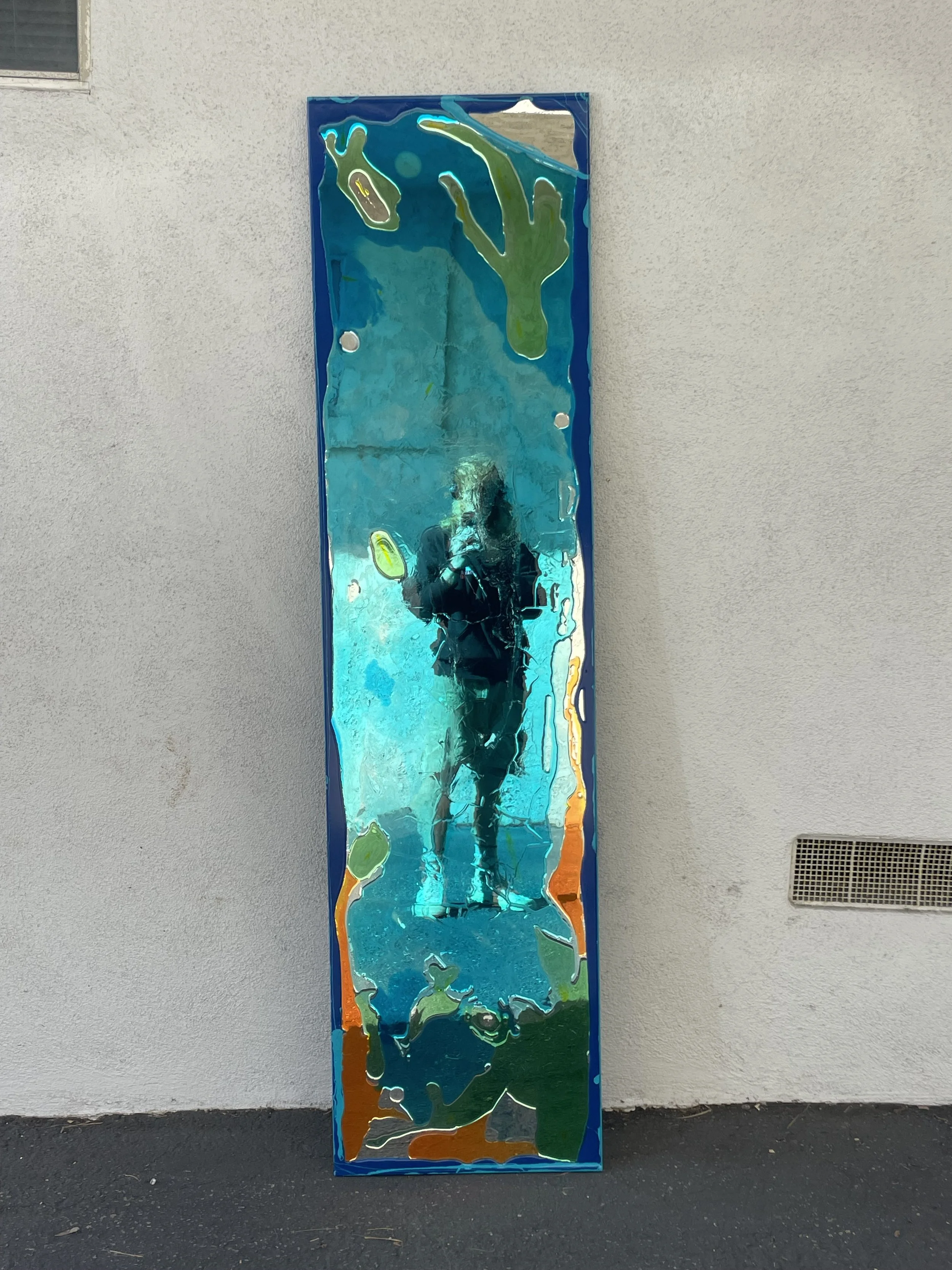 A tall, rectangular resin mirror with colorful abstract border, reflecting a person taking the photo, standing outdoors against a gray wall, with a vent near the bottom right.