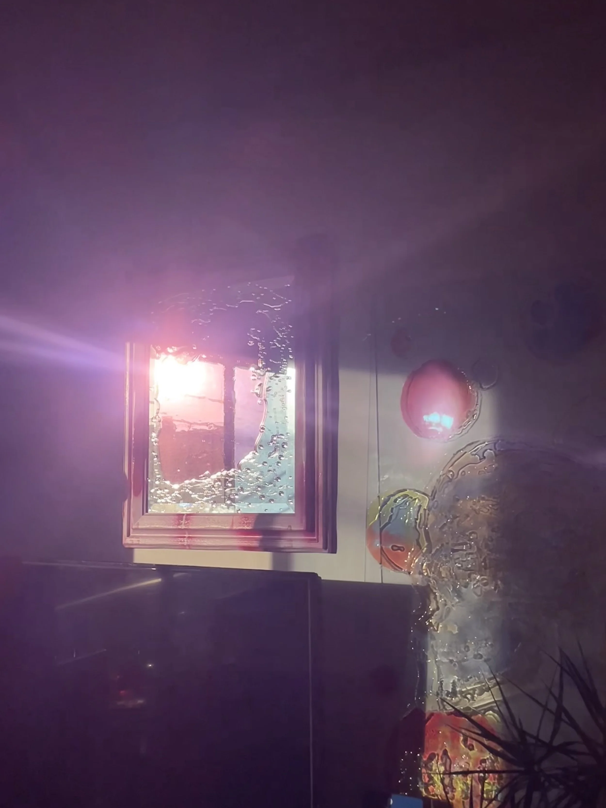 Indoor scene with a window letting in bright pinkish sunlight, several colorful resin balls on a table, a TV screen, and a plant in the foreground.