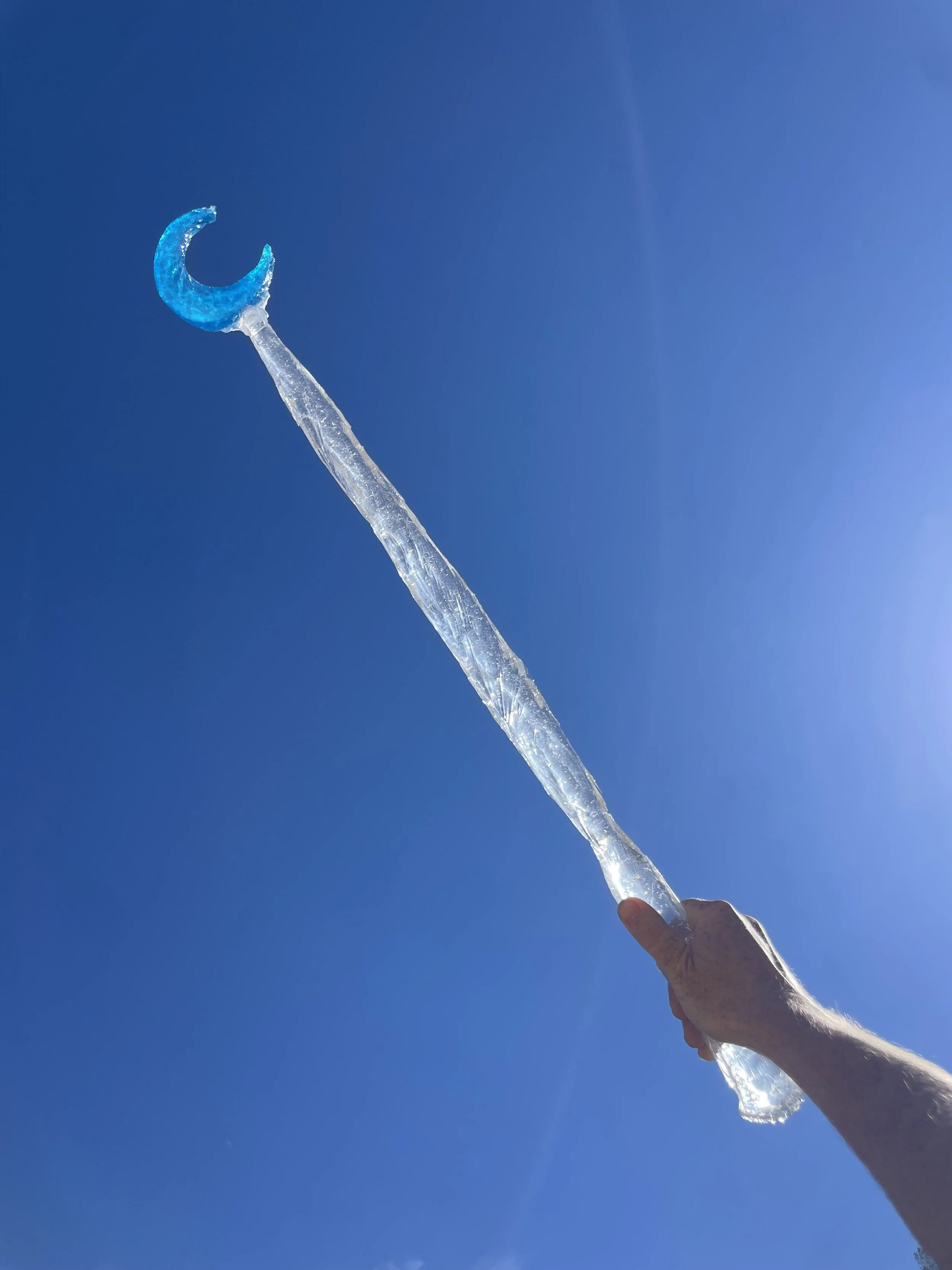 A person's hand holding a large, transparent, blue-tinted ice-like resin magic wand with a crescent moon shape at the top, against a clear blue sky.