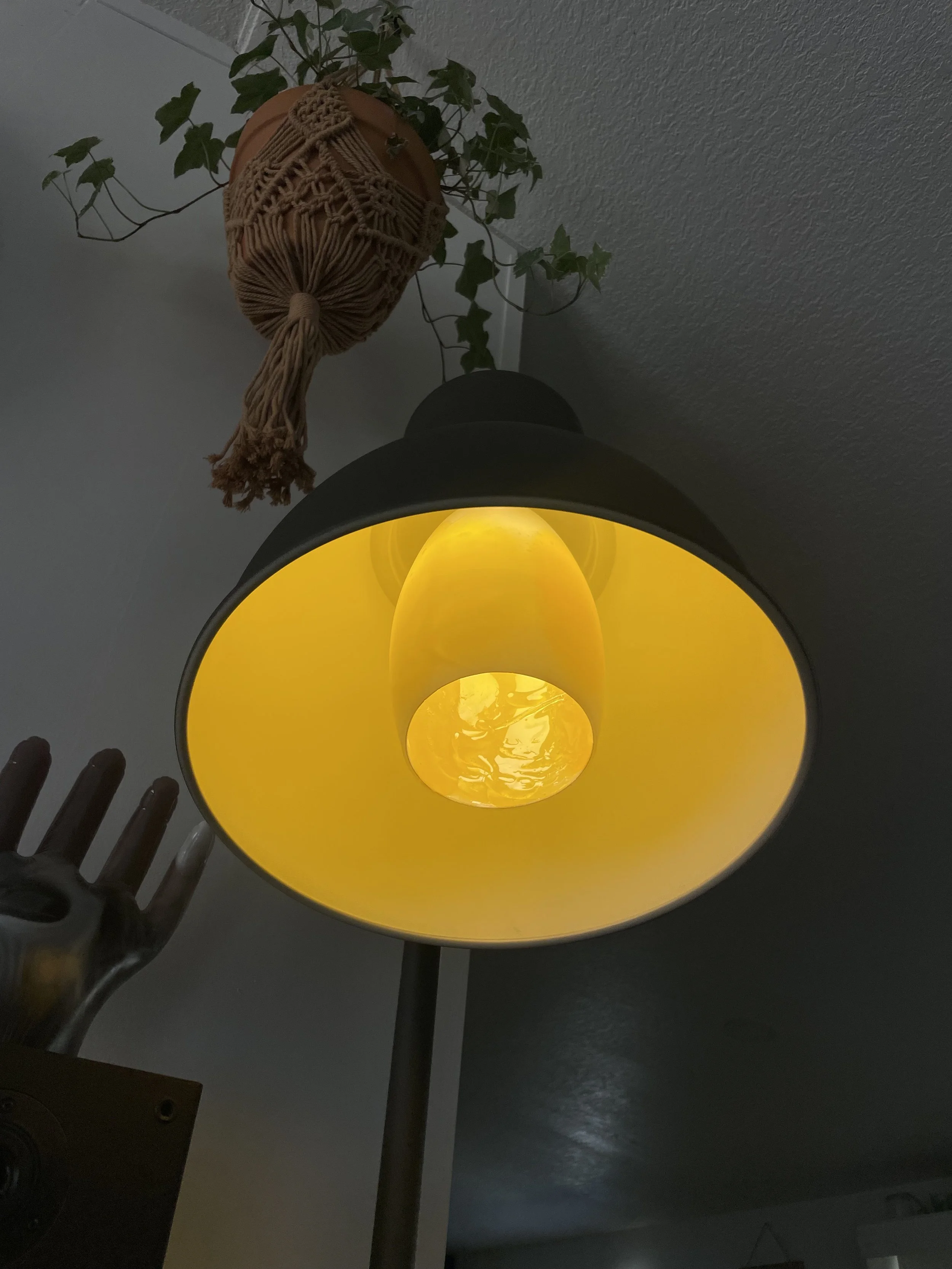 A dark gray floor lamp with a yellow interior resin lampshade, viewed from below, with a hand-shaped object and a plant in a hanging pot in the background.