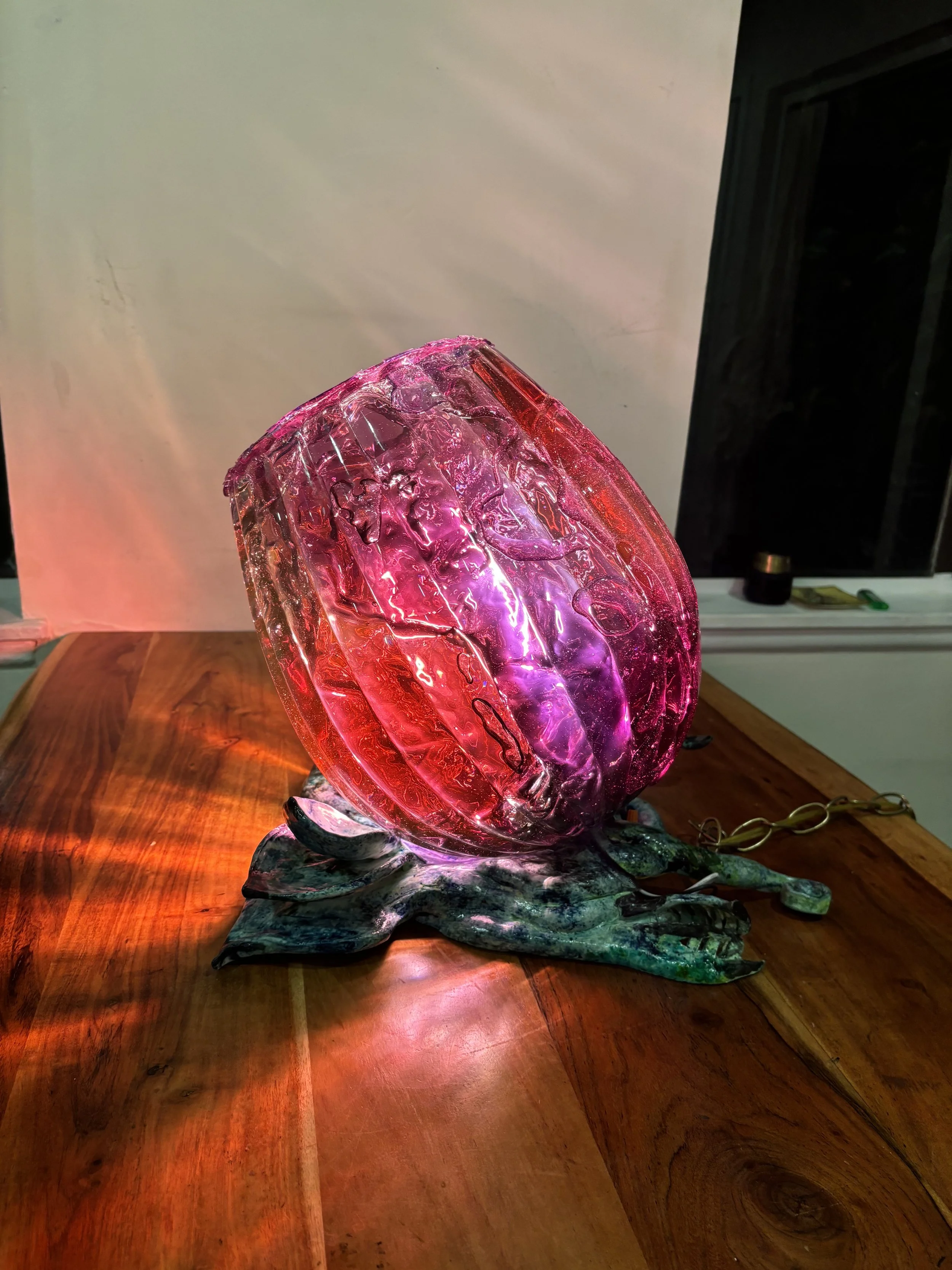 A decorative glass sculpture of a luminous pink and purple glass orb resting on a metal leaf-shaped base, placed on a wooden table.