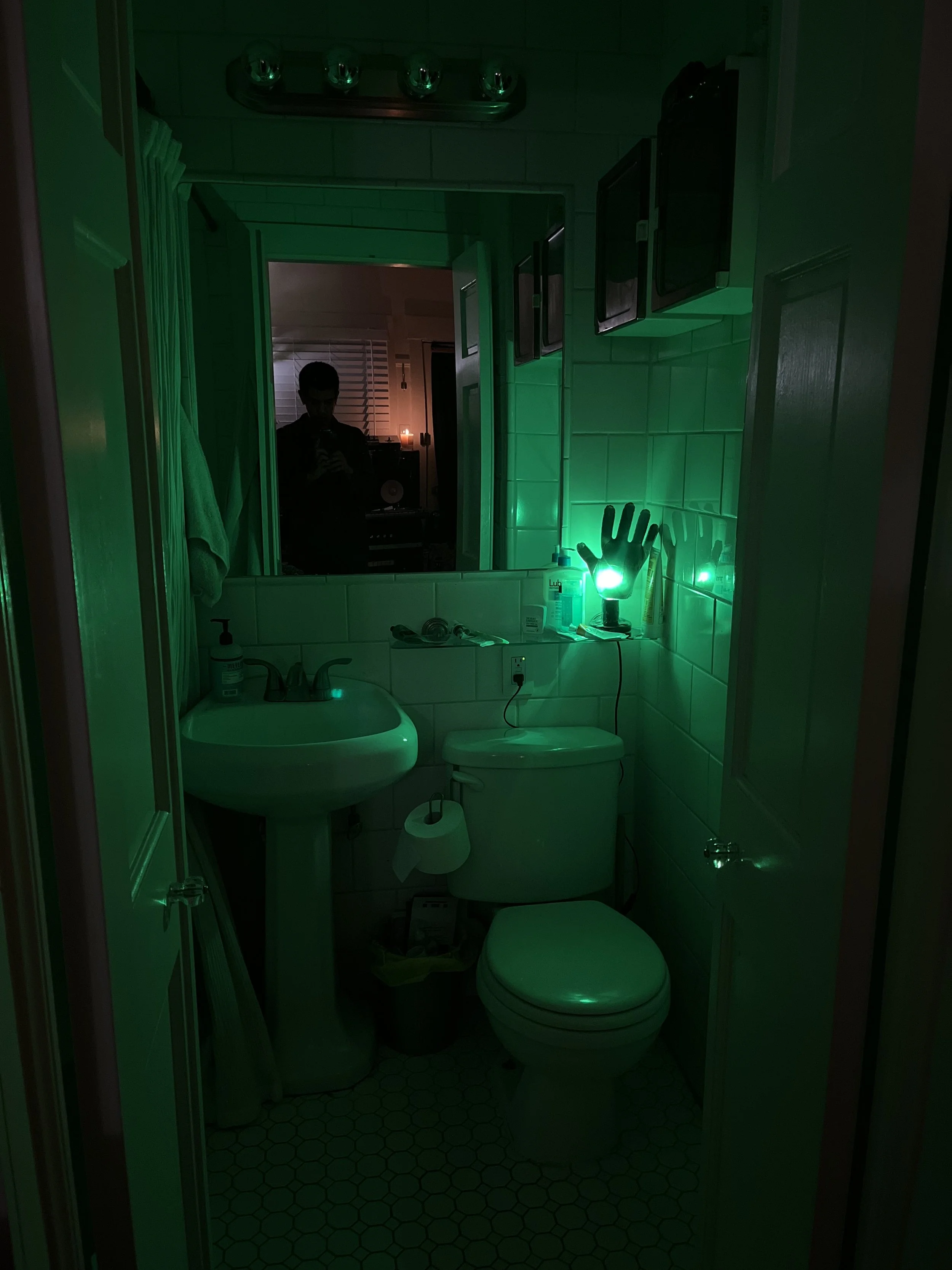 Bathroom illuminated by green light inside of a black resin hand, showing a sink, toilet, mirror, and various toiletries.
