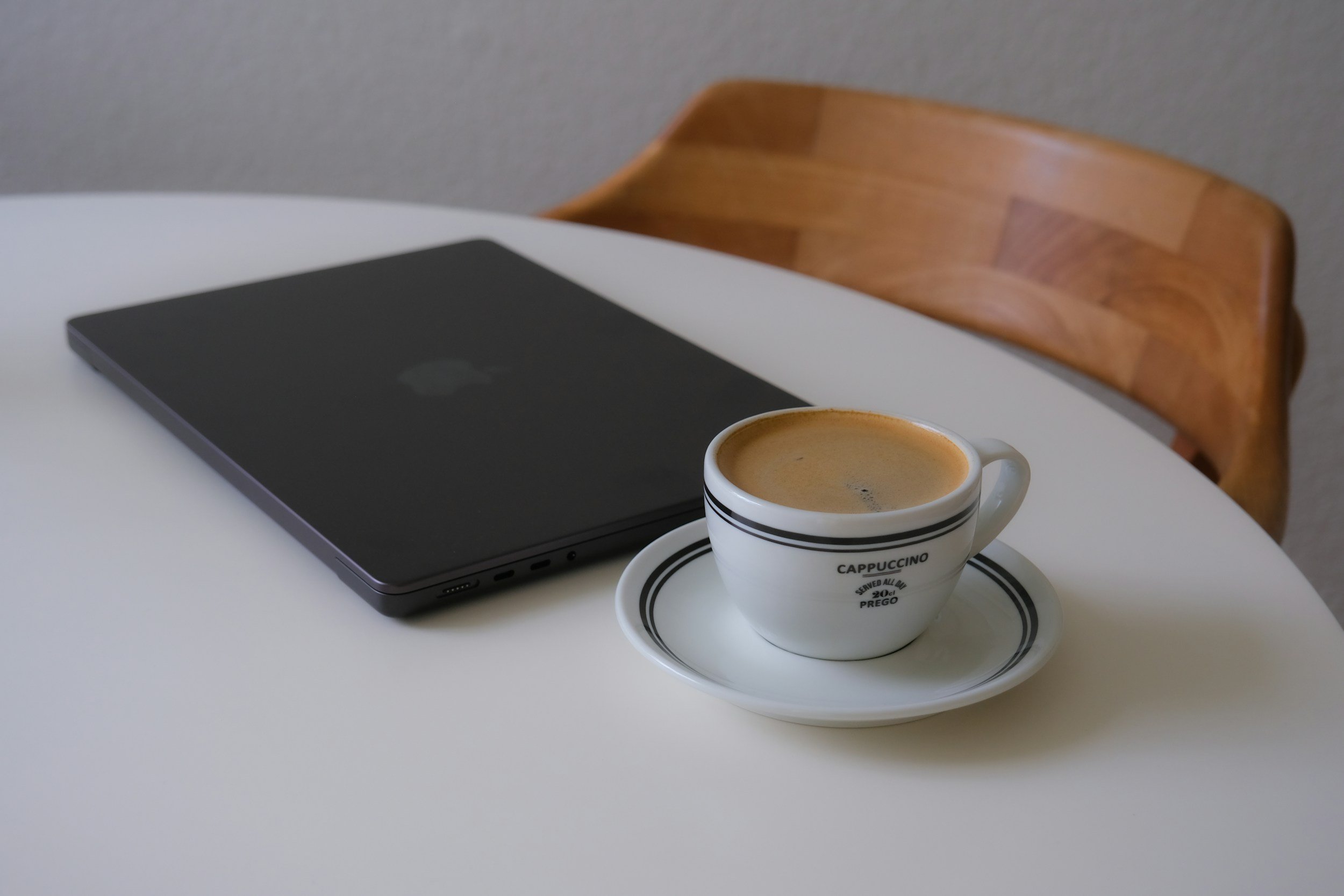 A closed laptop and a cup of cappuccino on a white table with a wooden chair in the background.