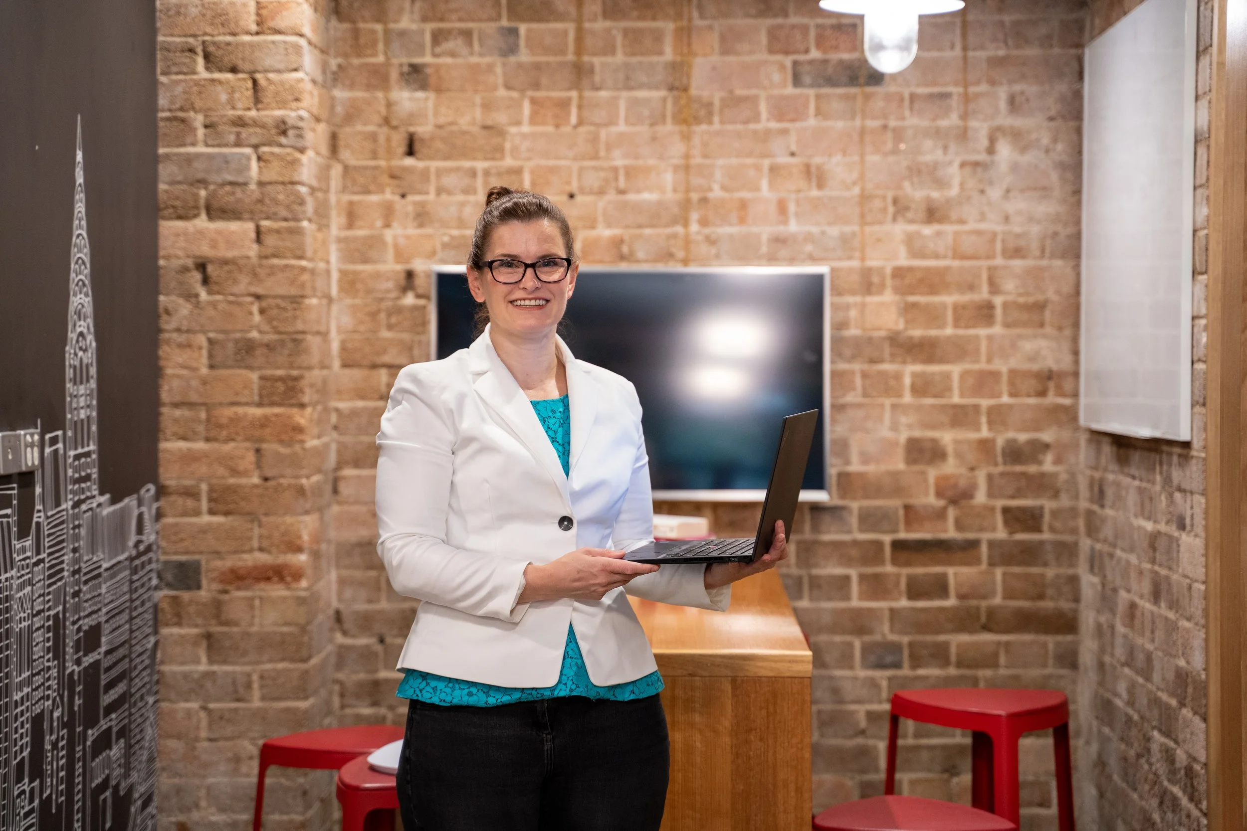 A woman with glasses and a ponytail, wearing a white blazer and teal top, smiling while holding a laptop in a room with exposed brick walls, a large monitor, and red stools.