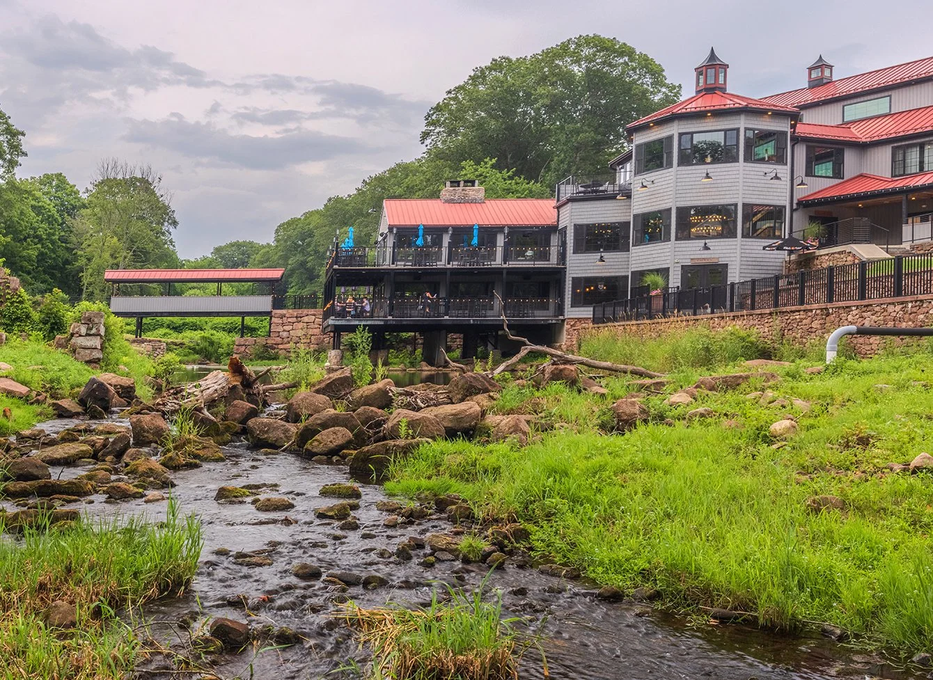 A river runs through a grassy area with rocks, leading to a large building with multiple levels and red roofs, set against a backdrop of trees and cloudy sky.