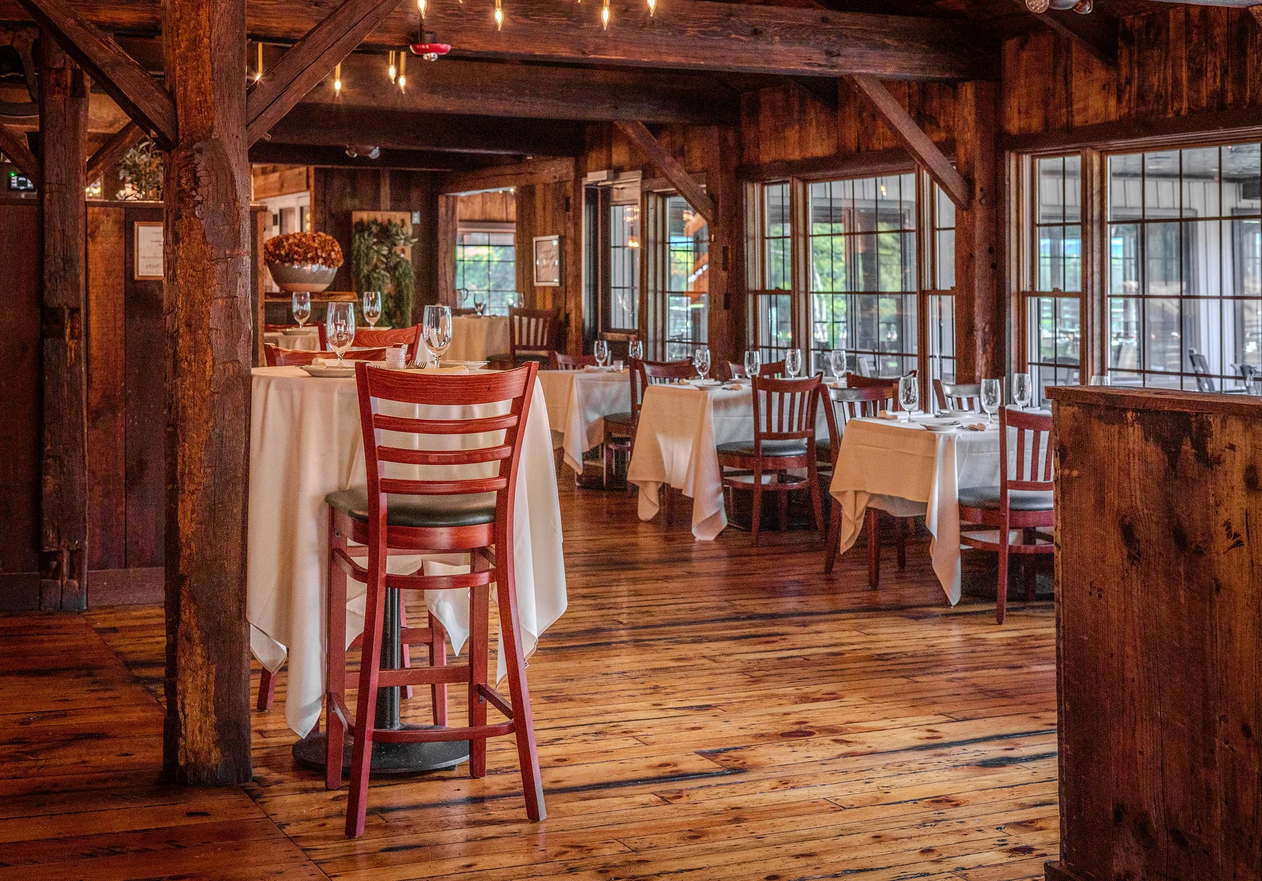 Elegant restaurant interior with wooden beams, hardwood floors, and tables set with white tablecloths, wine glasses, and silverware. Large windows let in natural light and offer outdoor views.