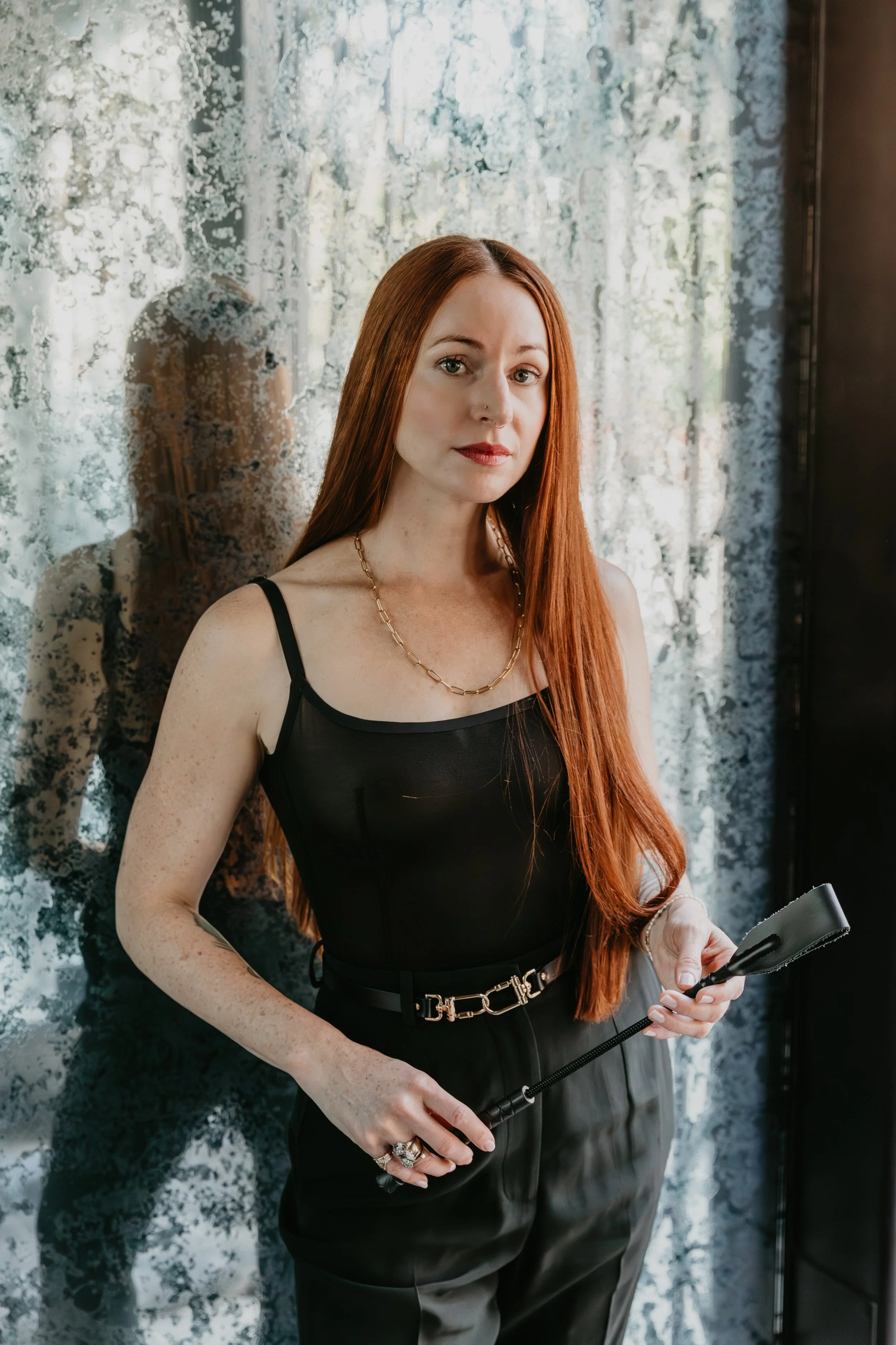 A woman with long red hair, wearing a black sleeveless top and black pants, holding a leather crop, standing next to a textured mirror or glass panel. She is a bdsm coach.