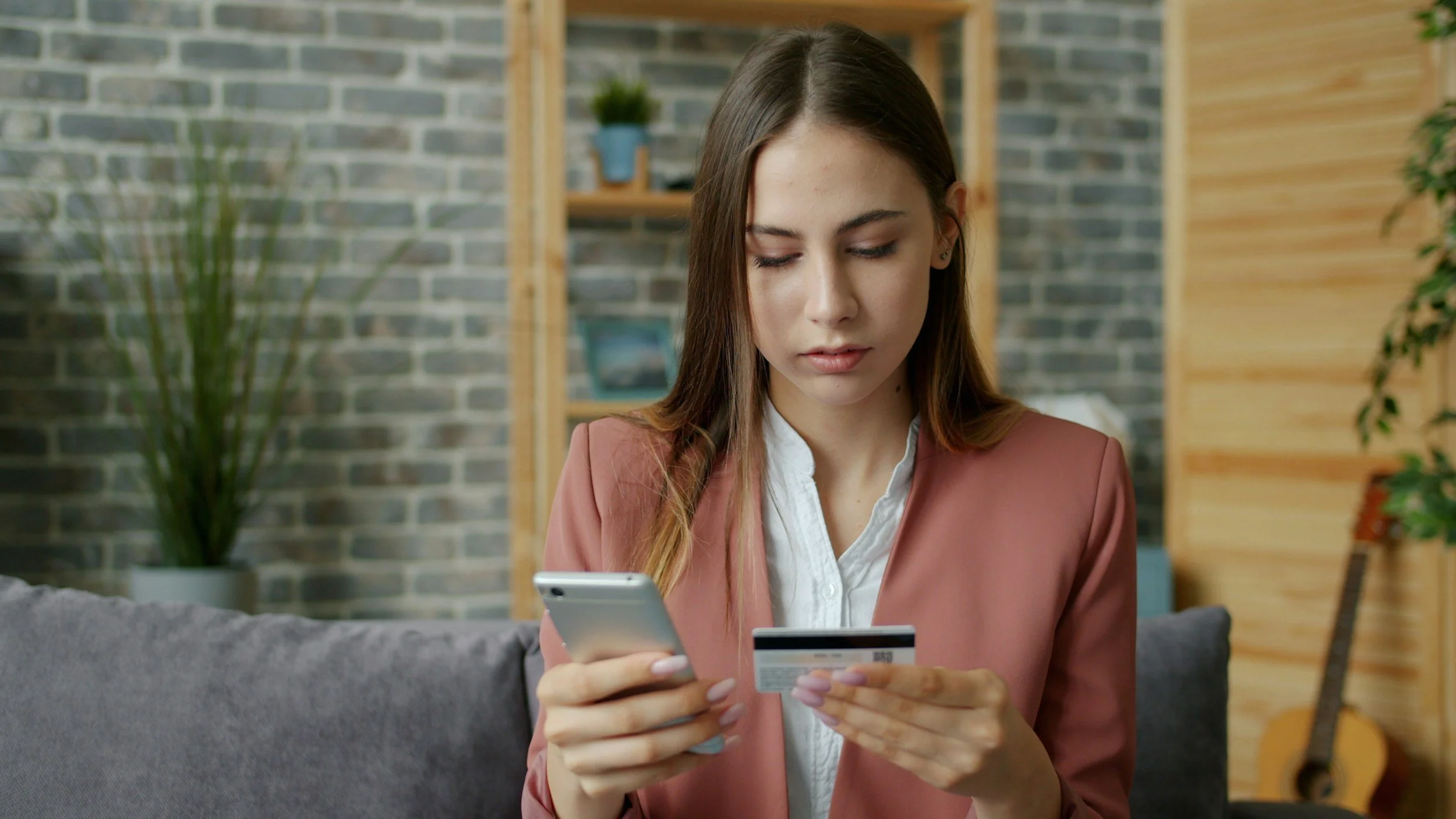A young woman in a pink blazer is looking at her credit card and smartphone while sitting on a gray couch in a modern living room.