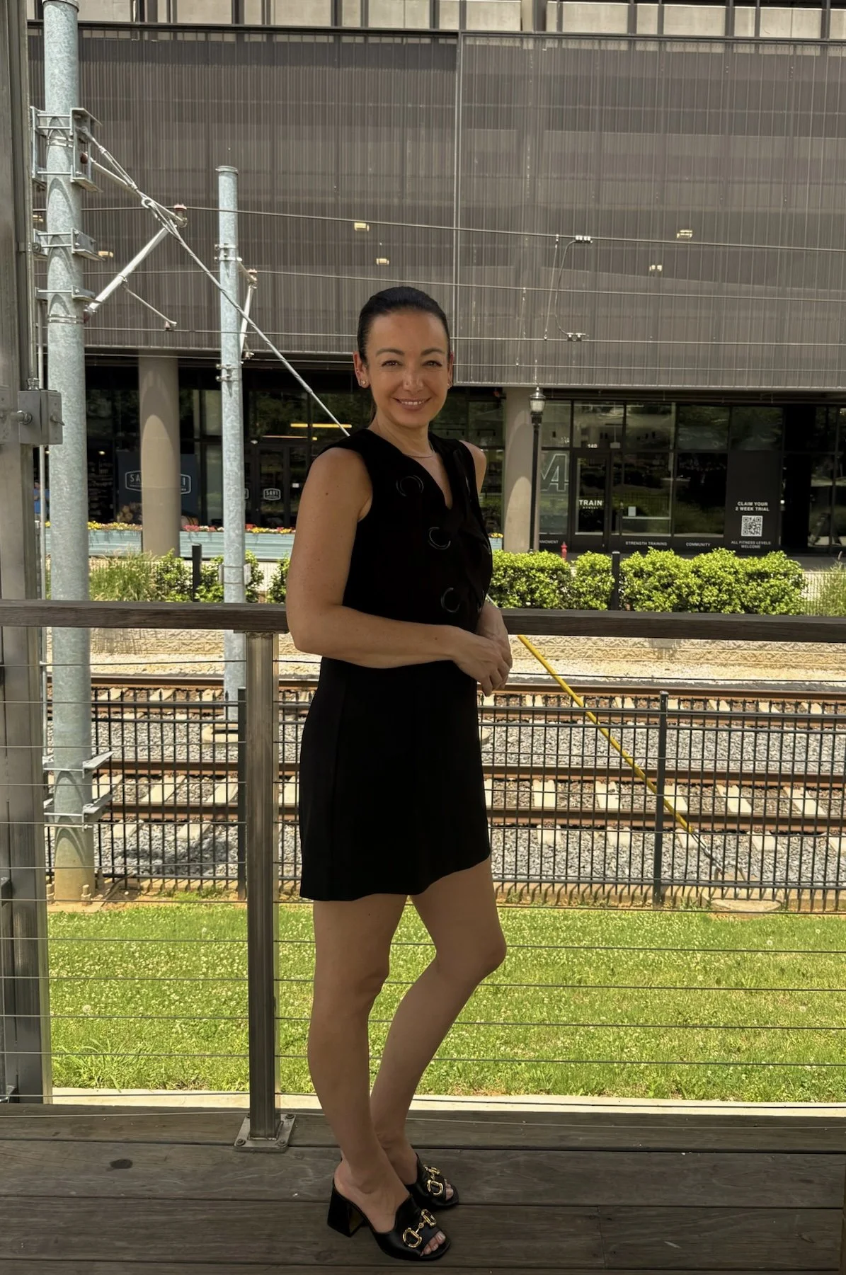 A woman standing on a wooden outdoor deck near train tracks, smiling, wearing a black sleeveless outfit and black heeled shoes, with a modern building in the background.