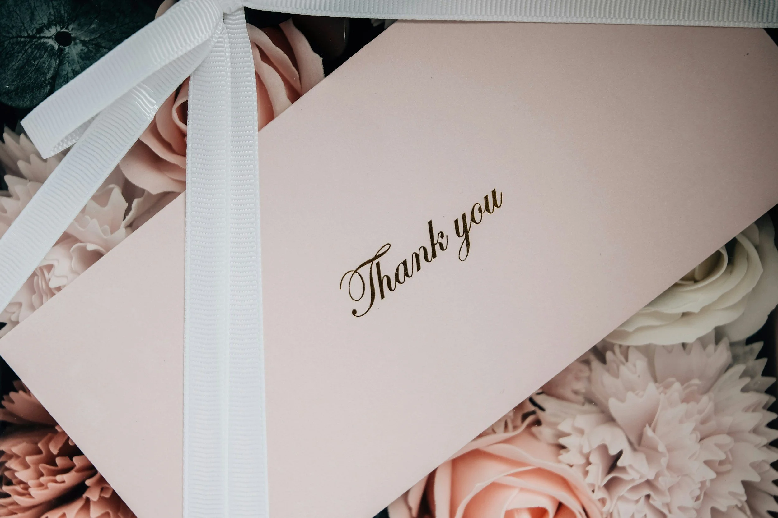 Close-up of a pink box with the words 'Thank you' written in gold, tied with a white ribbon, surrounded by pink and black paper flowers.