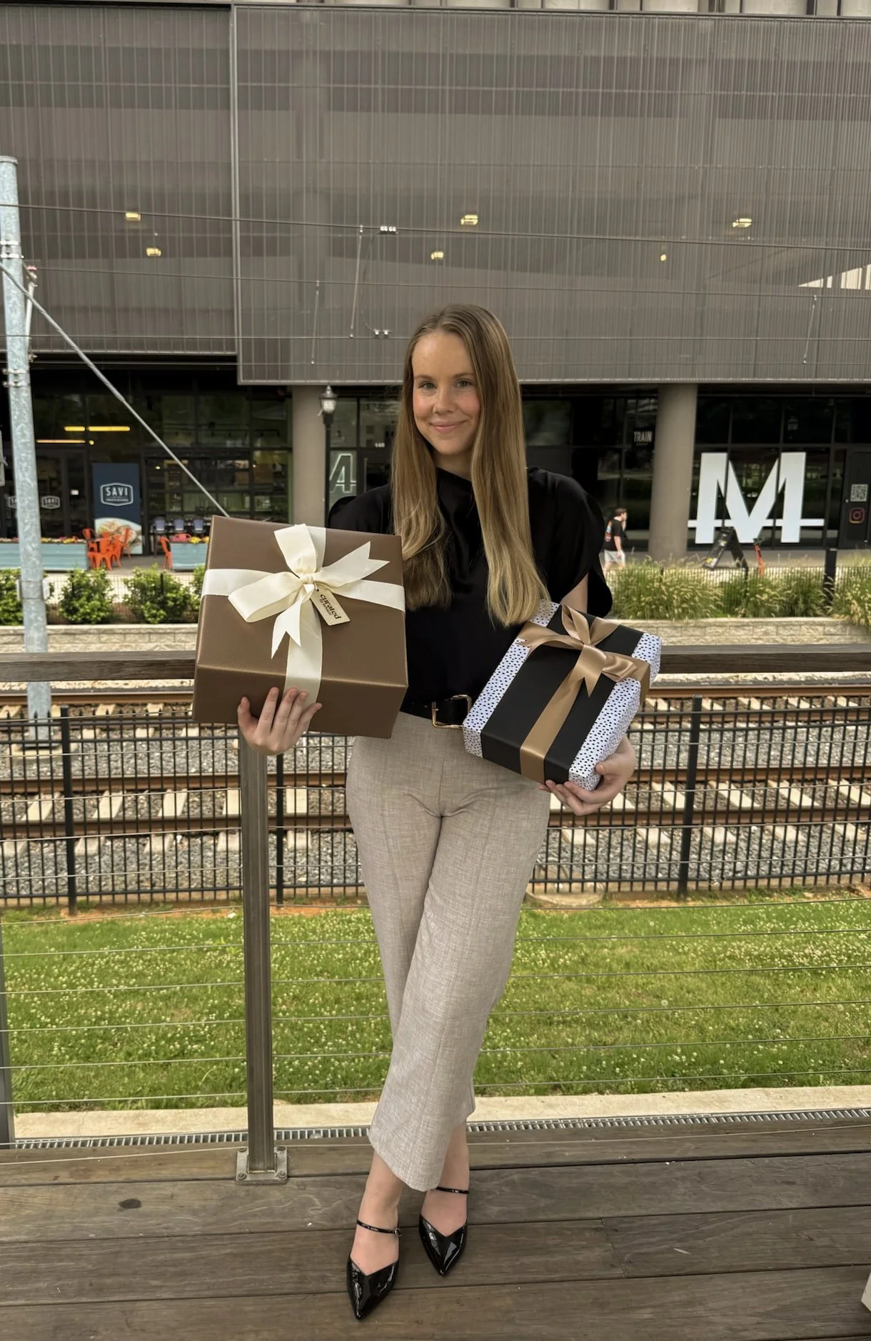 A woman with long light brown hair, wearing a black top and beige pants, stands outside holding two wrapped gift boxes, one with a white ribbon and the other with a brown ribbon. She is smiling and standing on a wooden deck with a metal railing, with train tracks and a modern building in the background.