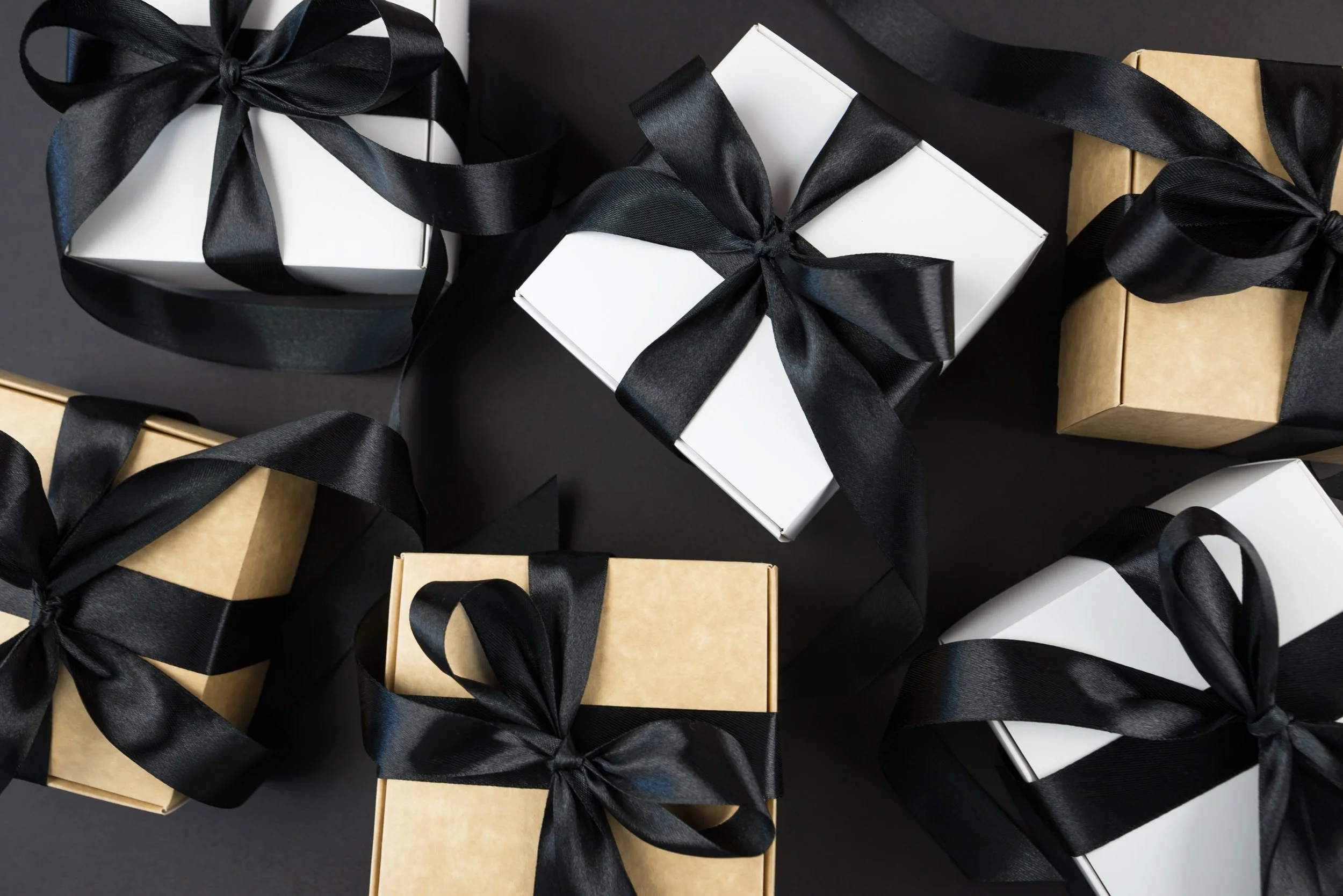 Multiple gift boxes wrapped in black, white, and gold paper with black satin ribbons on a black background.
