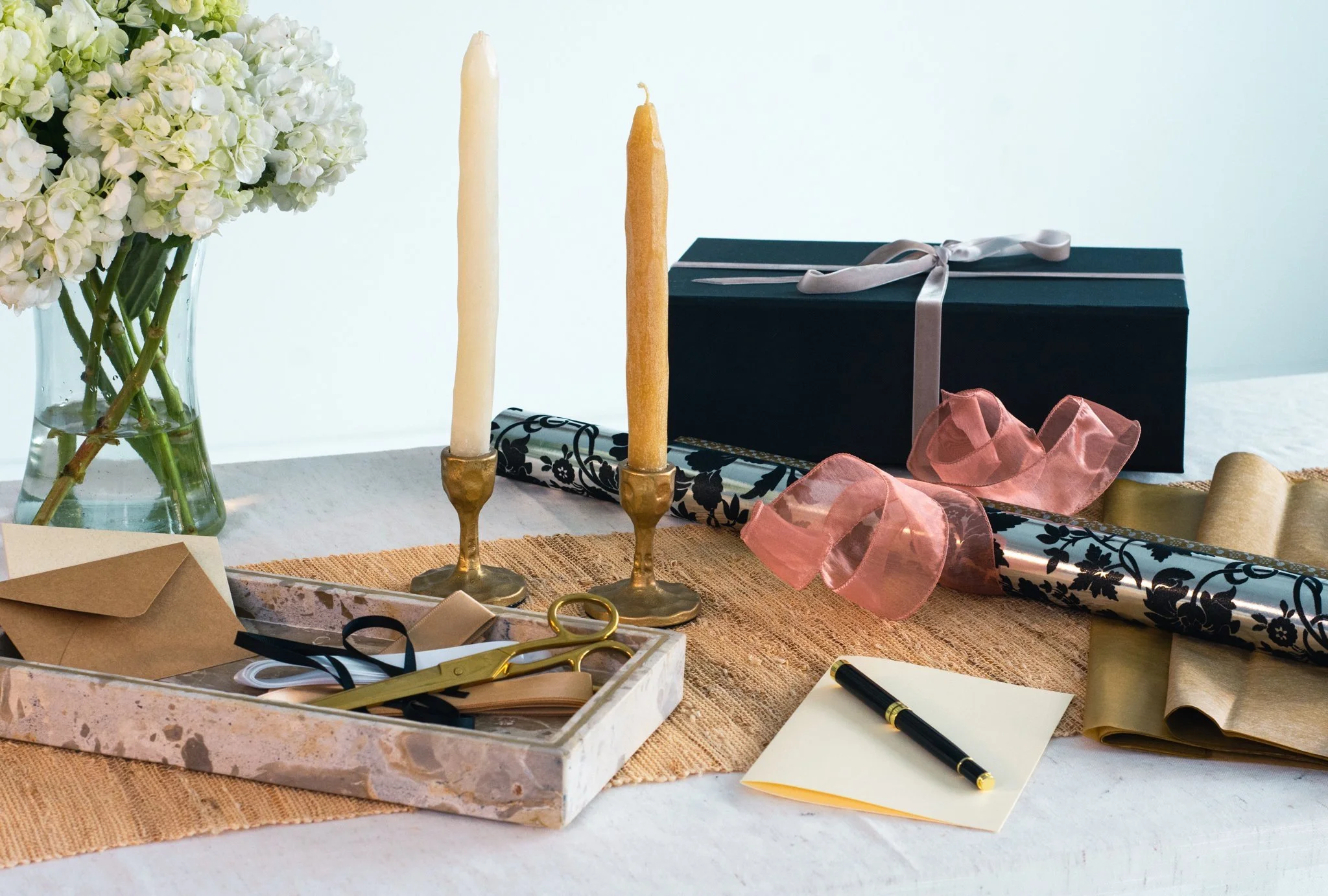 A table with white hydrangea flowers in a glass vase, two taper candles in gold holders, wrapping paper, scissors, ribbons, a marker, and a gift box.