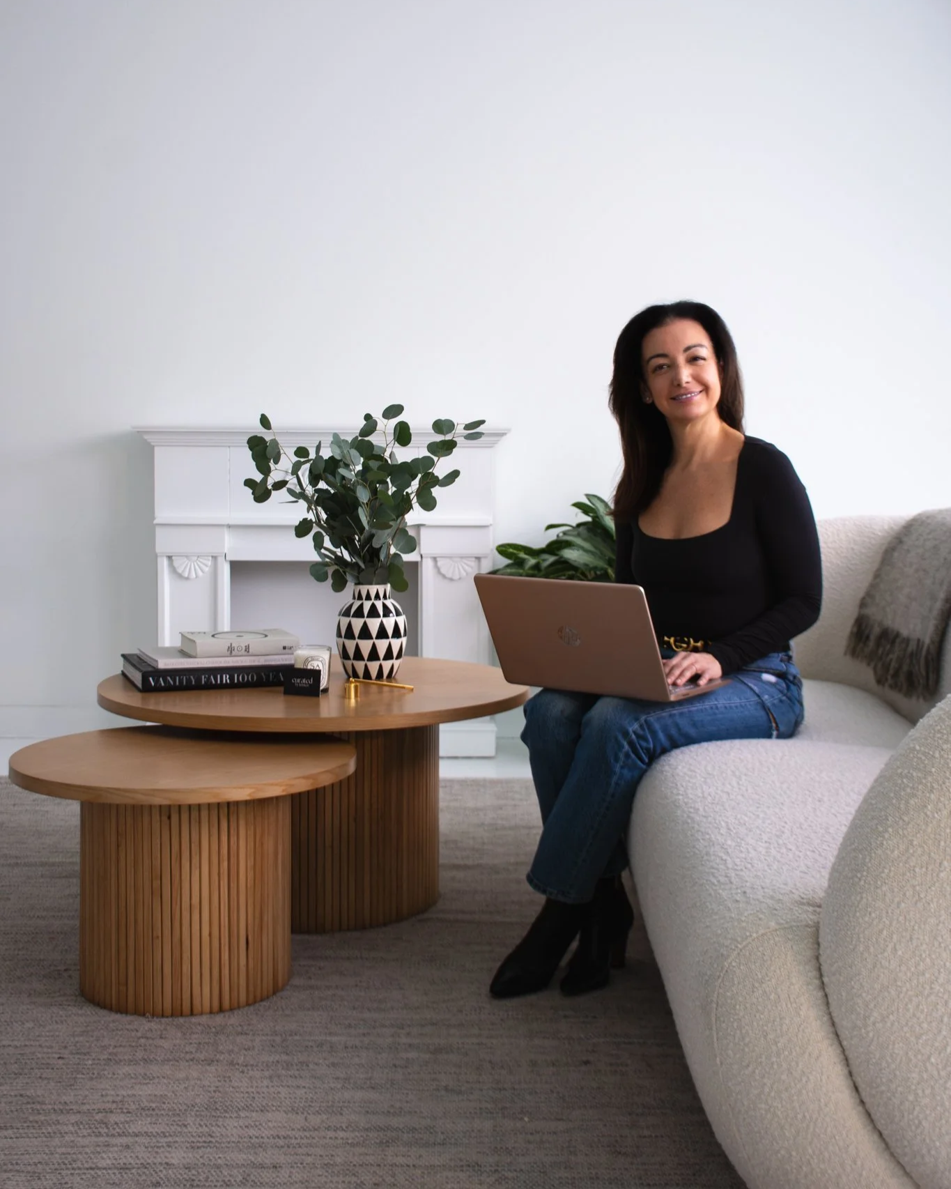 A woman with dark hair, wearing a black top and blue jeans, sitting on a white couch with a silver laptop on her lap, smiling in a modern living room with a round wooden coffee table, a plant, books, and a white fireplace mantel.
