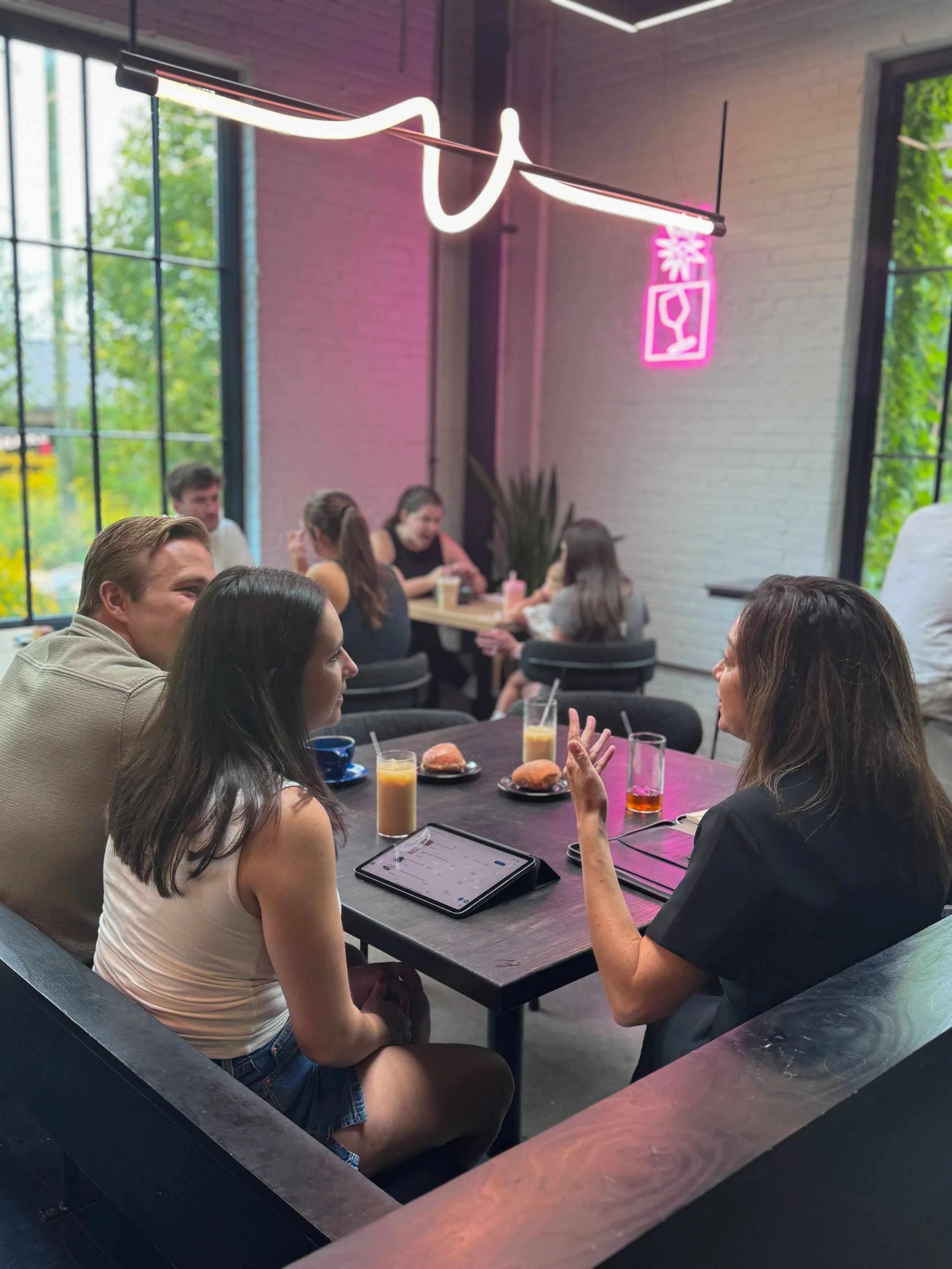 A group of people having a discussion at a long table in a modern, well-lit cafe or restaurant with large windows, pink neon signs, drinks, and pastries on the table.