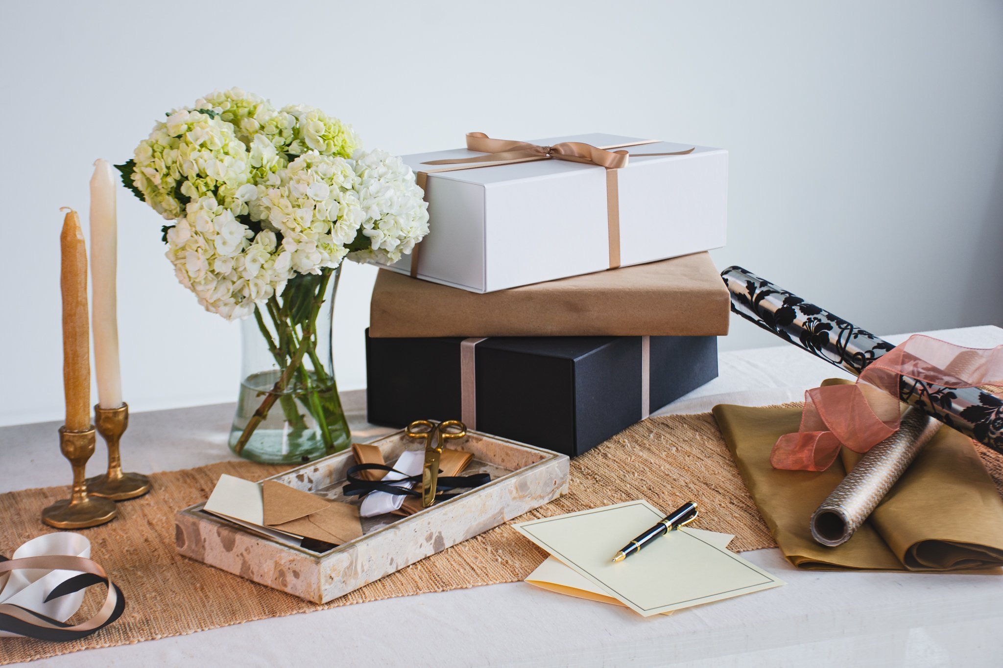 A collection of gift boxes on a table, including one white box with a beige ribbon, floral arrangements, candles, ribbons, and writing supplies.