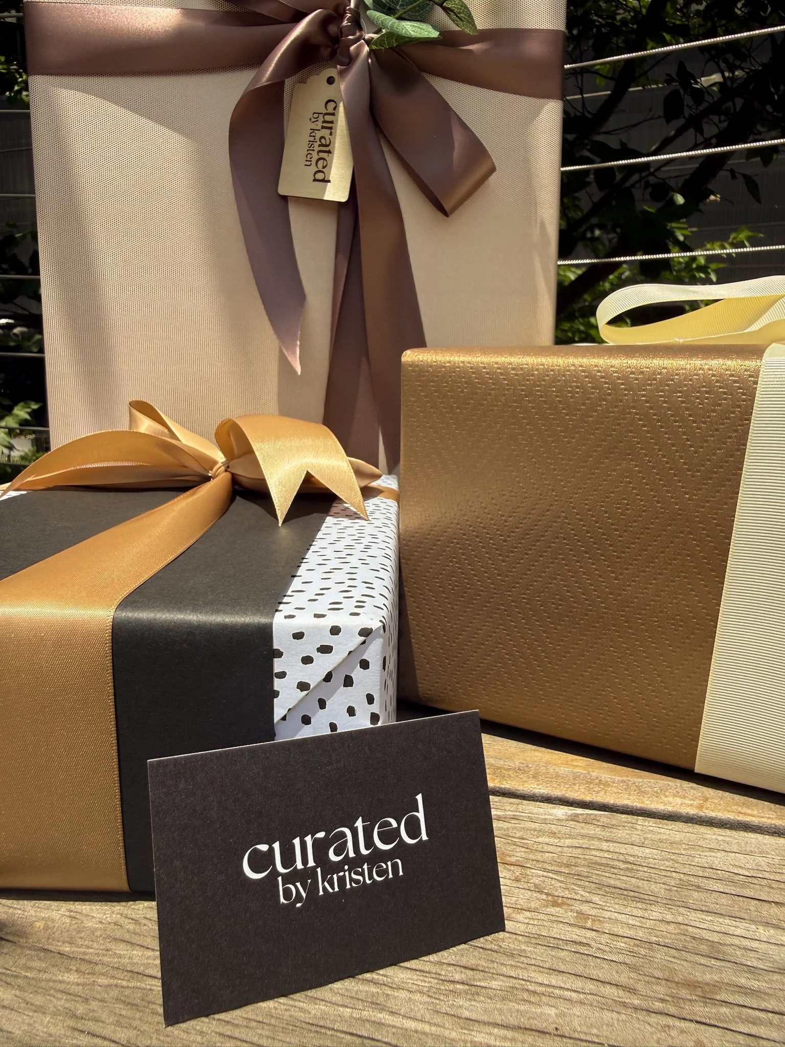 A beautifully wrapped gift with a beige and gold wrapping paper, tied with a large gold ribbon. There is a black tag with white text that reads 'curated by kristen'. The gift is placed on a wooden surface with other partially visible wrapped presents nearby.