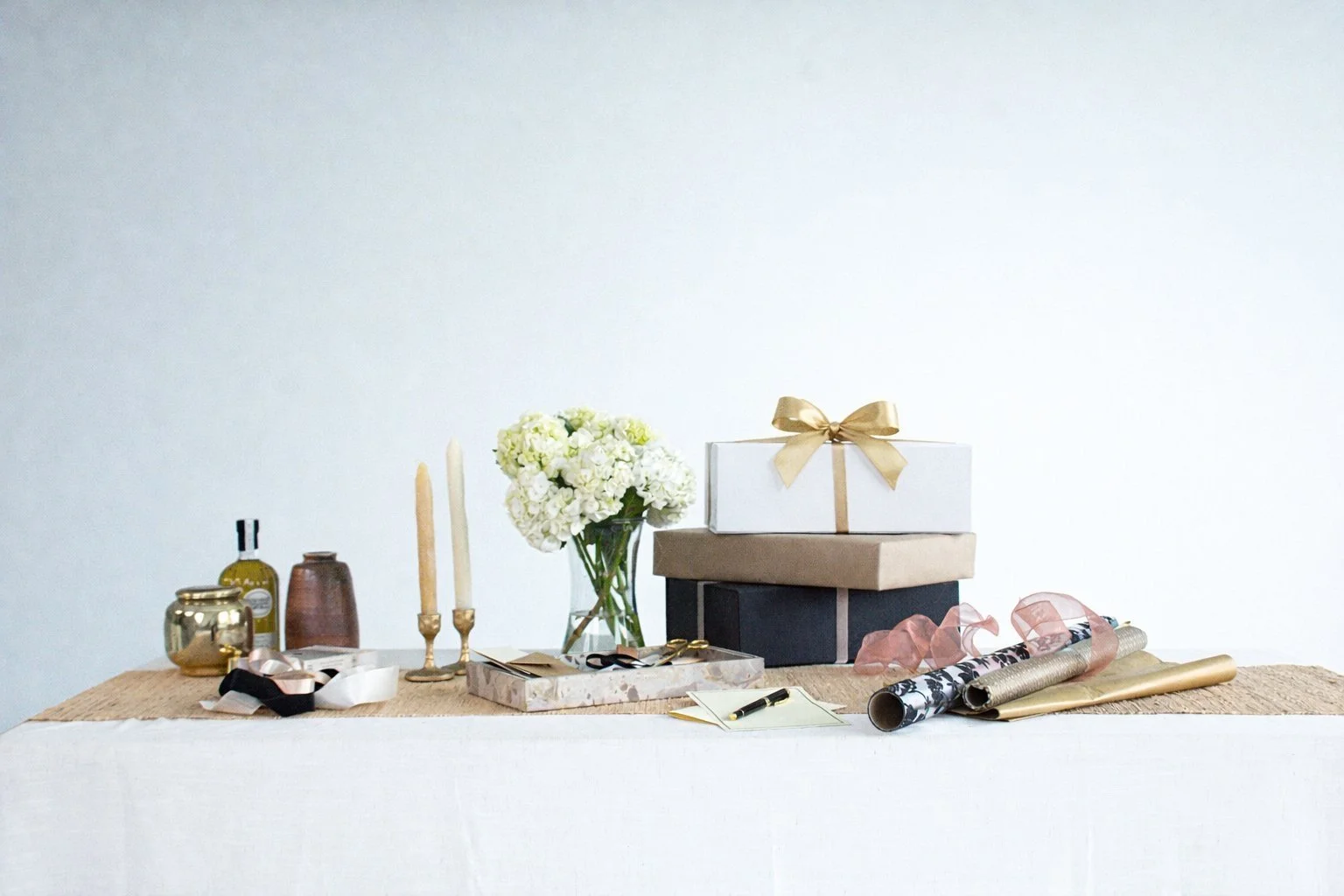 Table with wrapped gifts, a vase of white flowers, candles, ribbons, and small decorative items, set against a plain white wall.