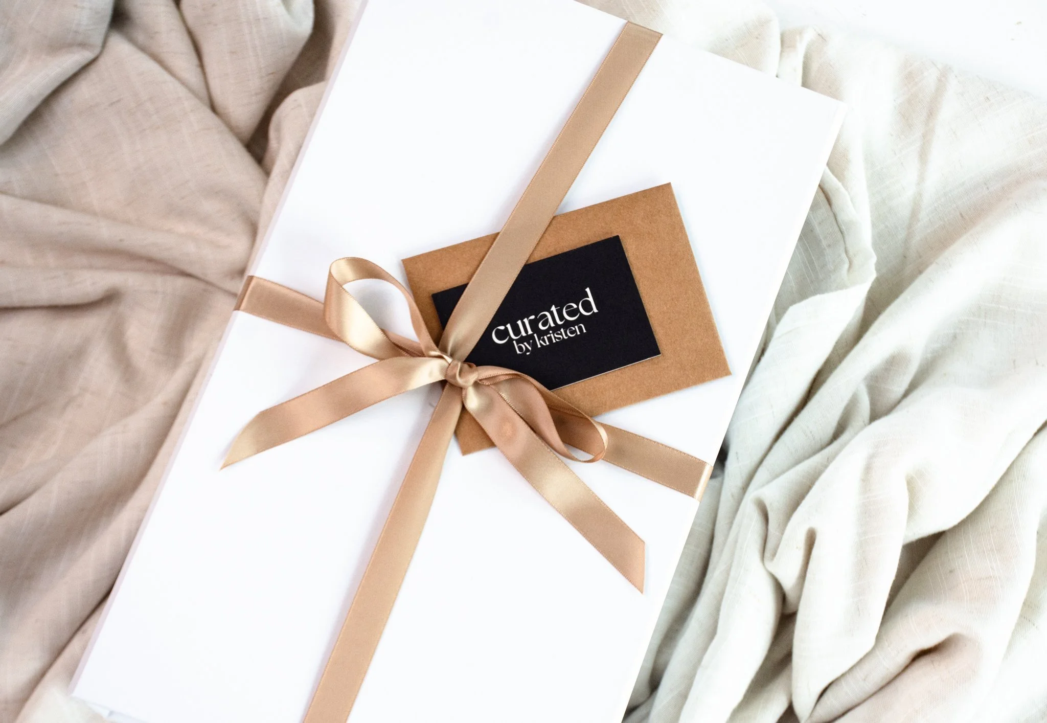 A white rectangular gift box with a tan satin ribbon tied in a bow, sitting on a beige and cream fabric surface. The box has a black label with white text that reads 'curated by kristen.'