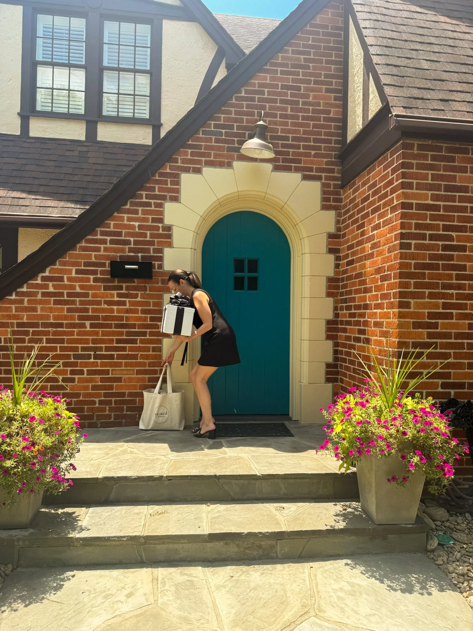 A woman in a black dress and high heels is cleaning the front porch of a brick house with a teal door, holding a white tote bag with a black strap. The porch has two large flower pots with pink and purple flowers on either side of the steps. The house has a black lantern-style light fixture above the door and a black mailbox to the left of the door. The house has a red brick exterior with some cream-colored trim around the windows and door.