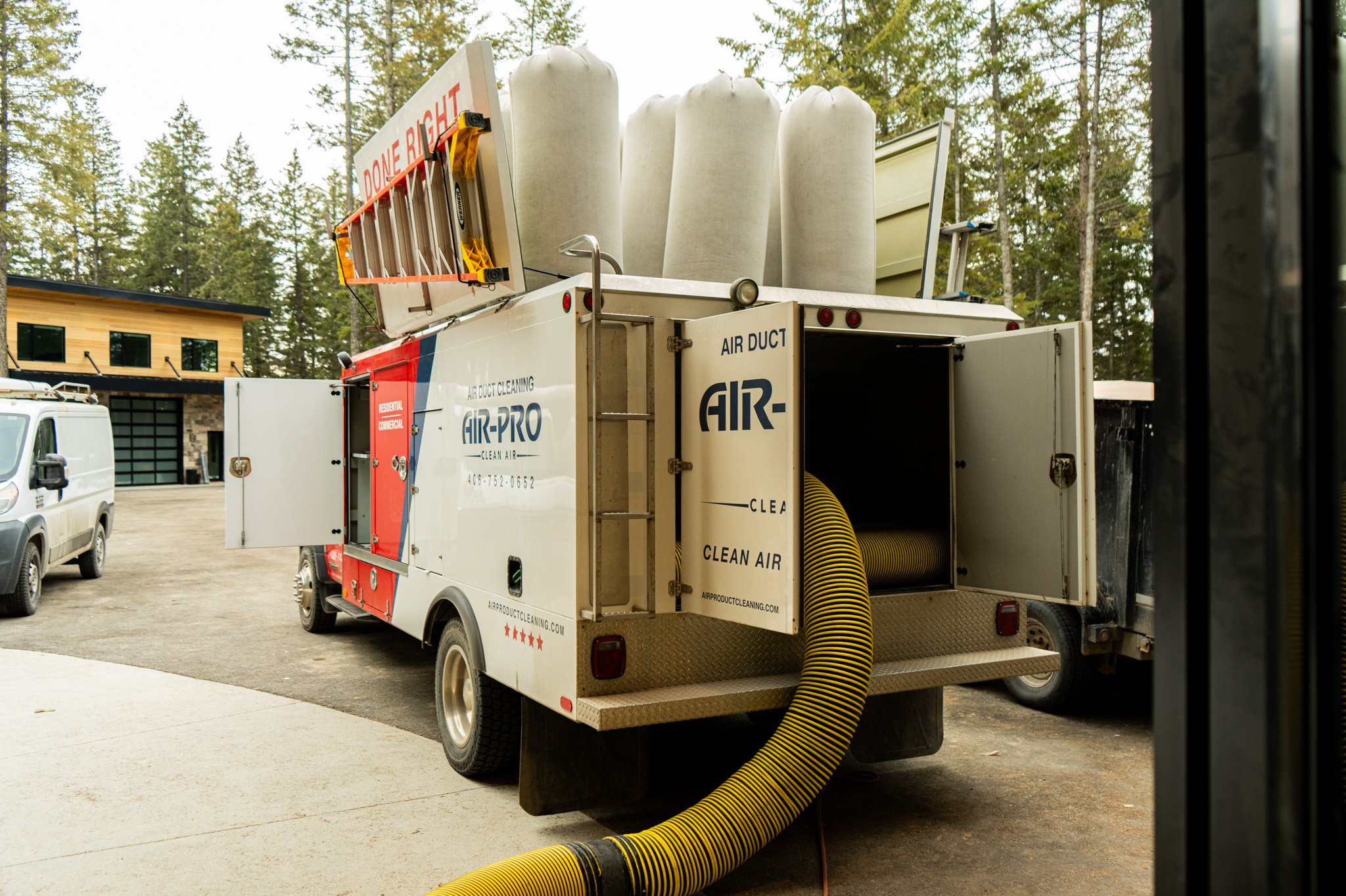 A mobile air duct cleaning truck with large white ducts on top and an open compartment with a yellow flexible hose attached