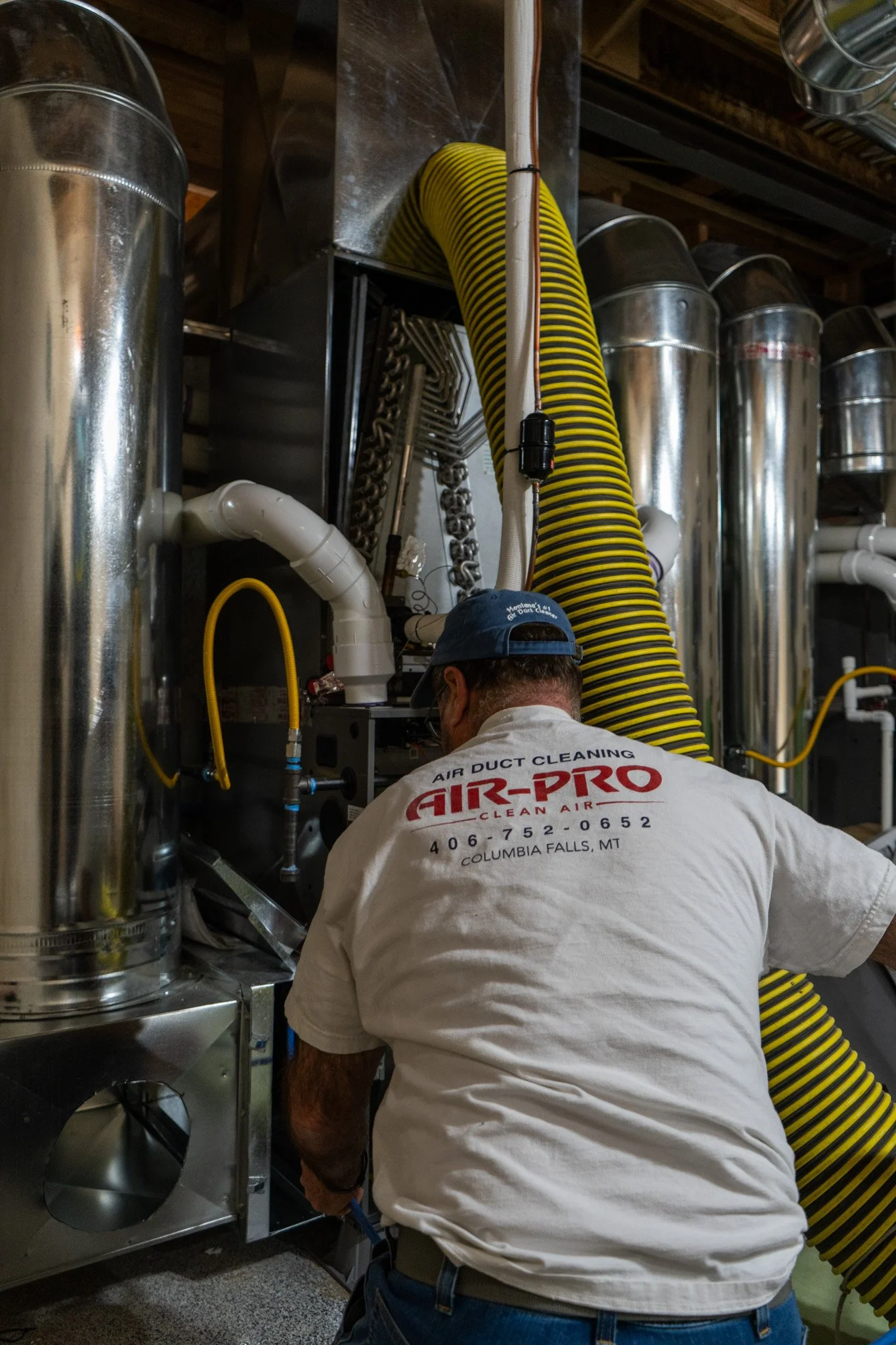 A man wearing a white shirt with 'Air-Pro' written on the back, working on air duct cleaning equipment in an industrial setting with large metal ducts and yellow flexible hose.