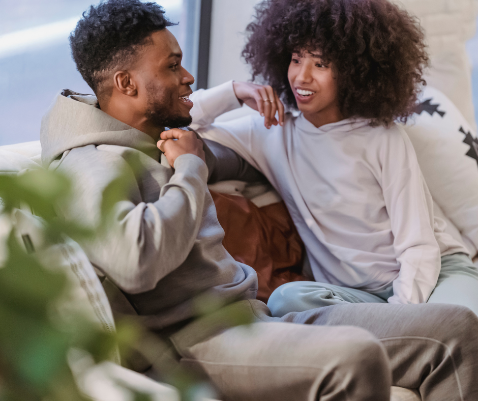 A young man and woman sitting on a couch, smiling and engaging in conversation indoors.