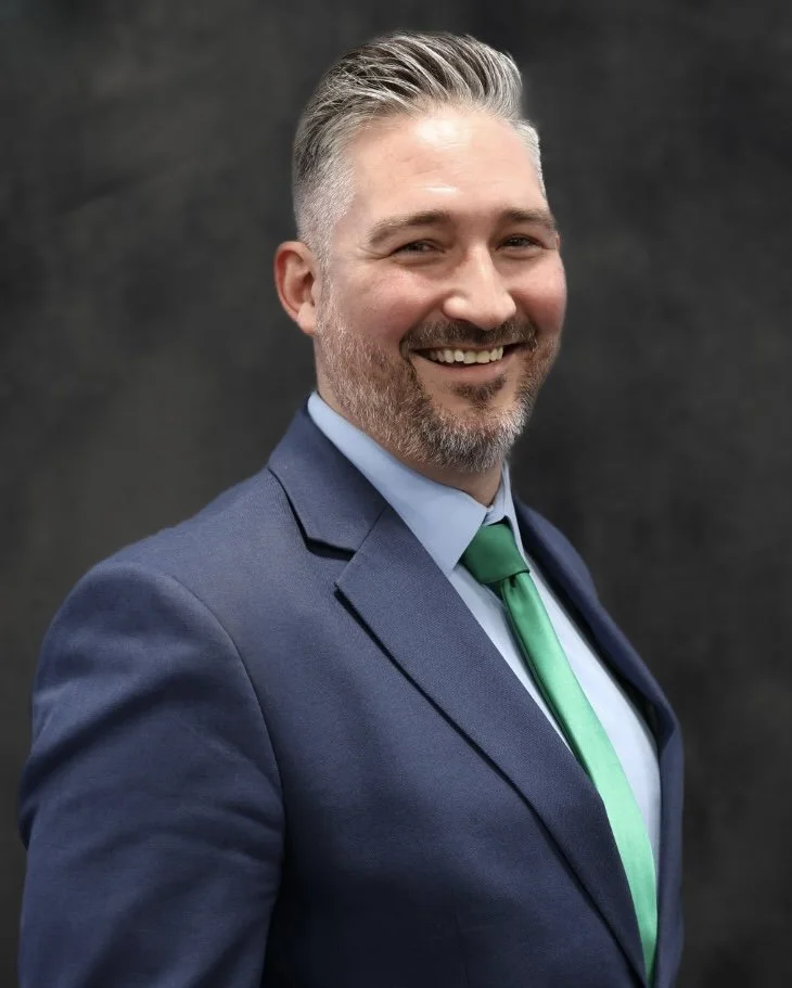A smiling man with gray hair and a beard, wearing a navy blue suit, light blue shirt, and green tie, standing against a dark background.