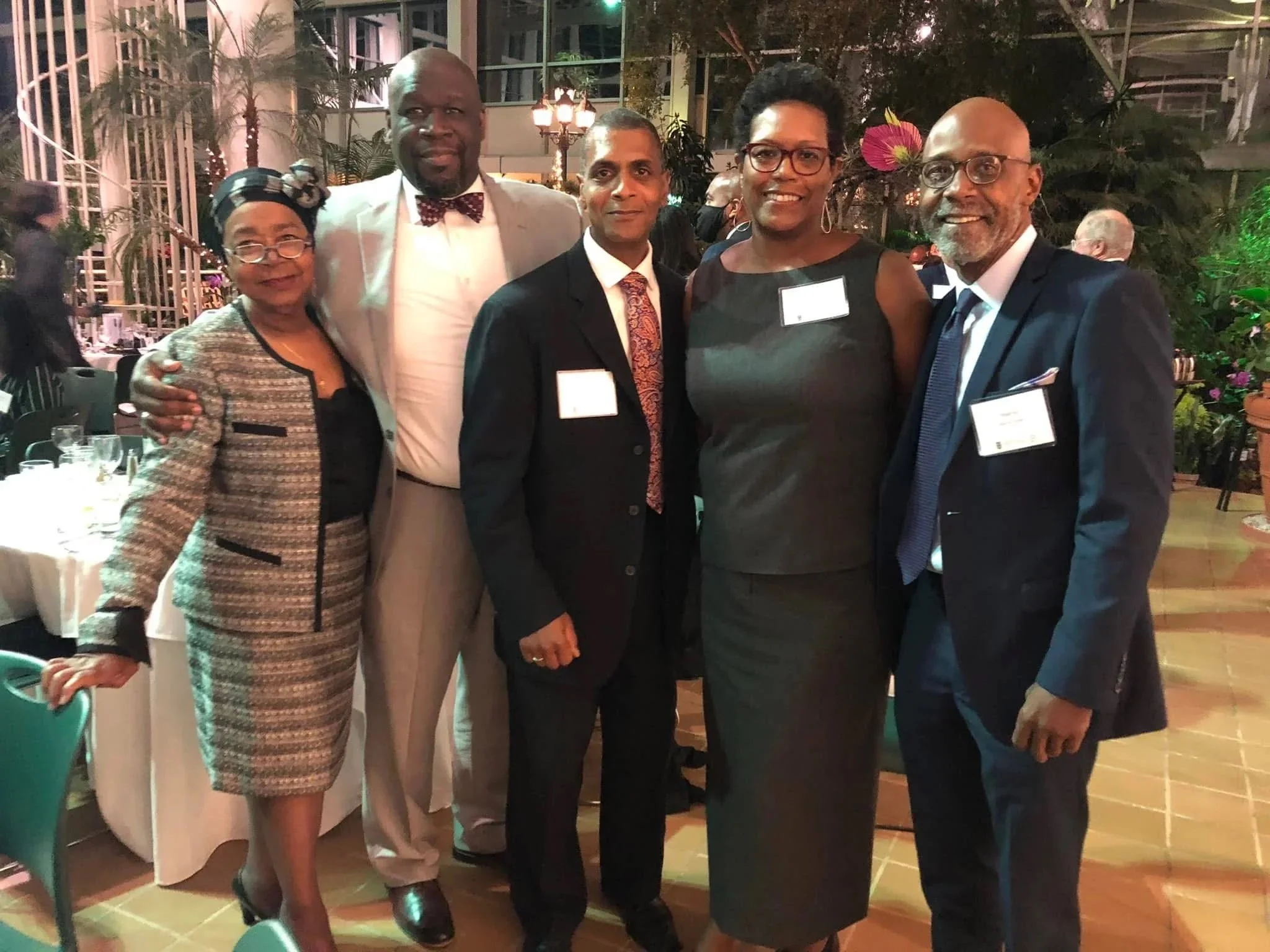 Group of five people dressed in formal business attire at a social event, standing indoors with plants and tables in the background.