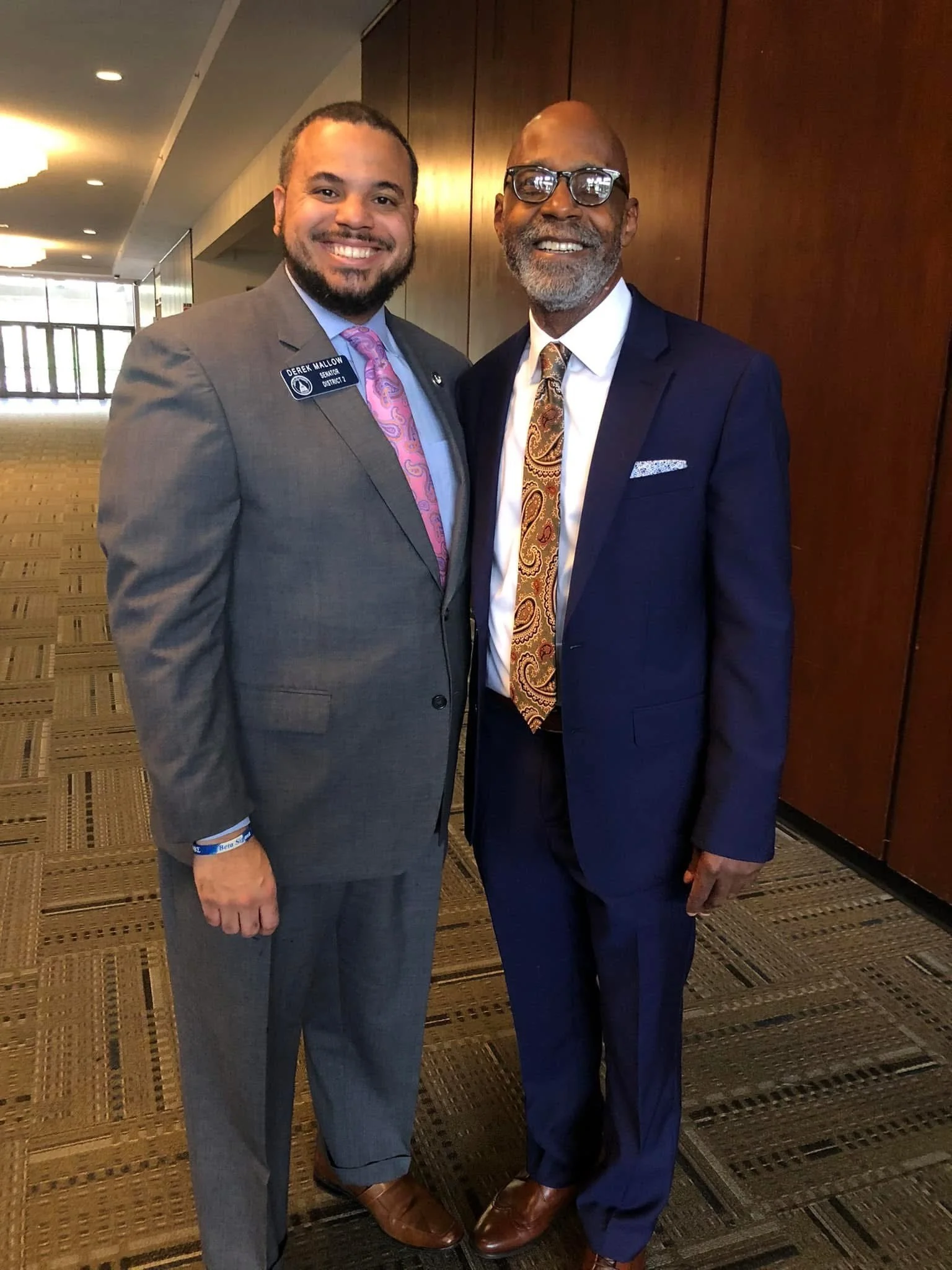 Two men in suits smiling and standing close together indoors, one wearing glasses and both dressed formally with ties.