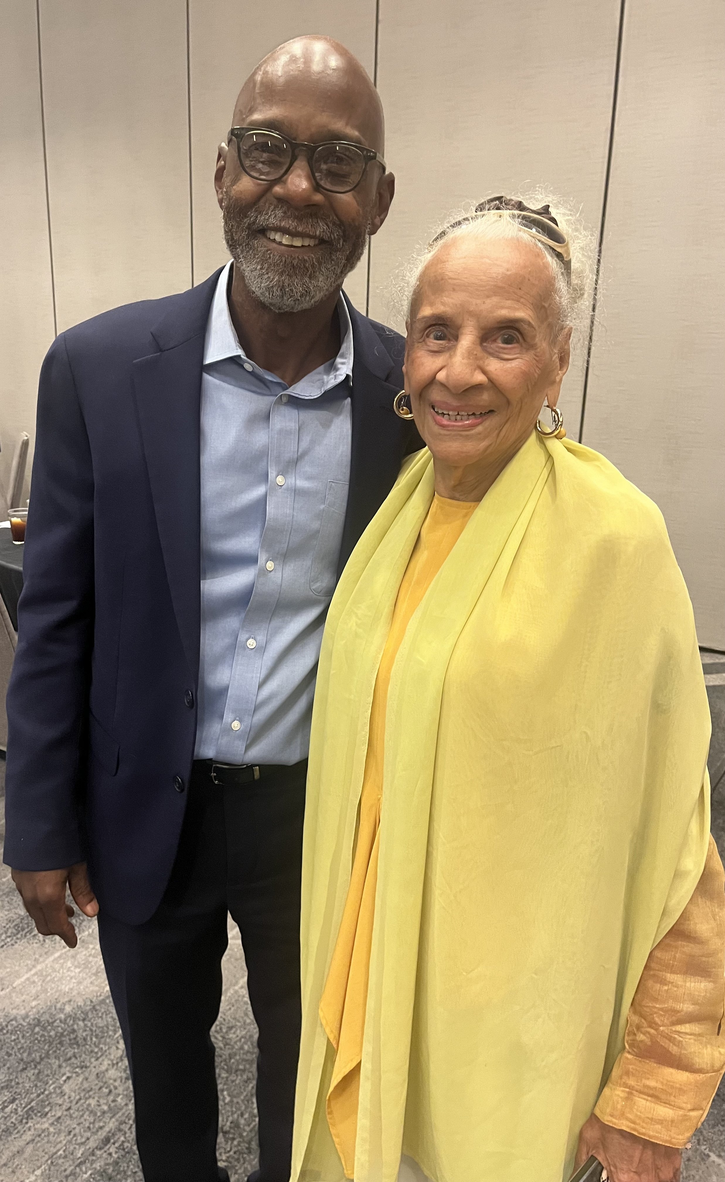 A man and an elderly woman standing together indoors, smiling at the camera. The man is wearing a navy blue suit, light blue shirt, and glasses. The woman is wearing a yellow shawl and earrings.