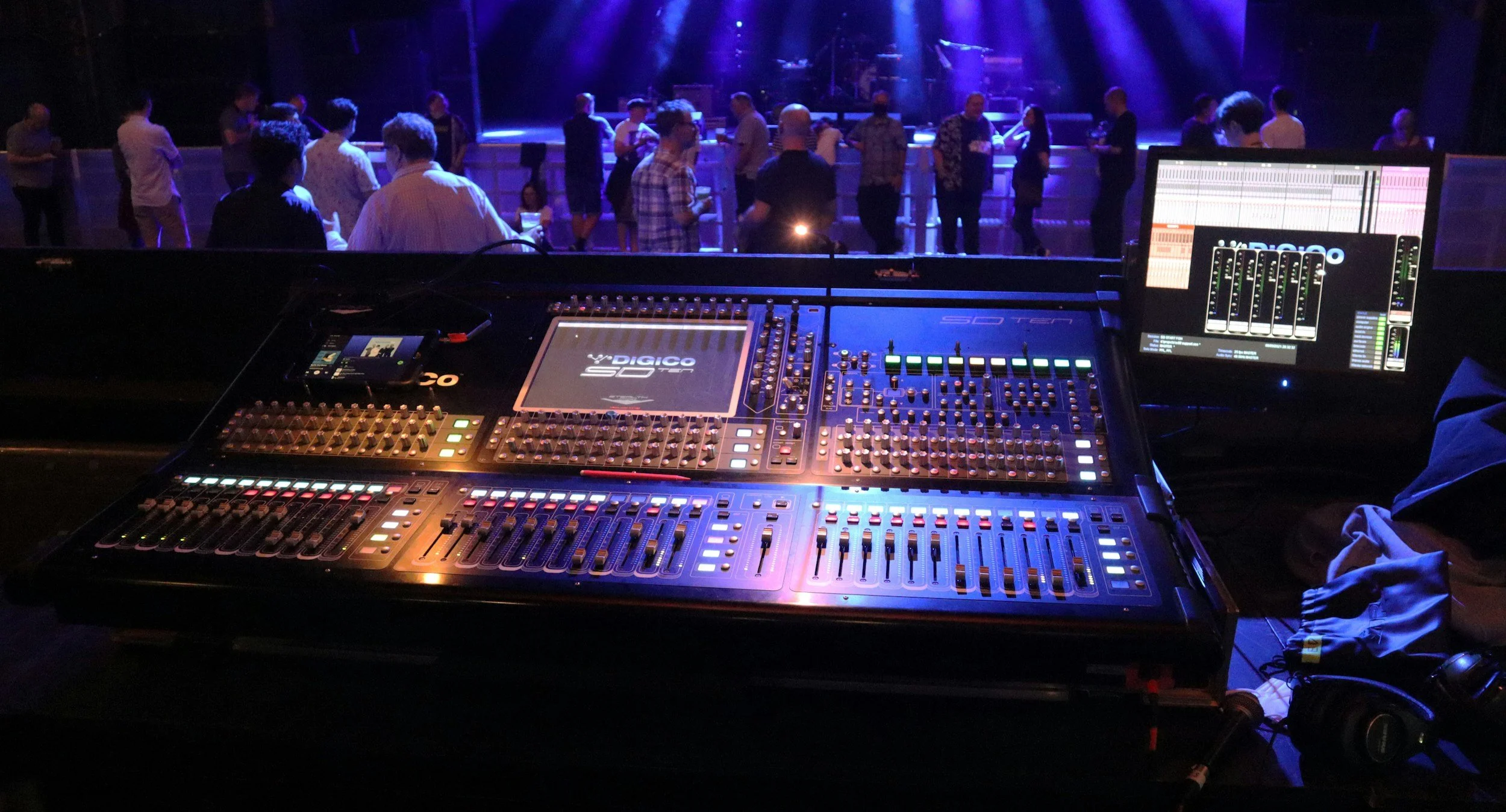 A view from the DJ's booth overlooking a dance floor with people socializing and dancing under purple and blue stage lights at a nightclub or concert venue.