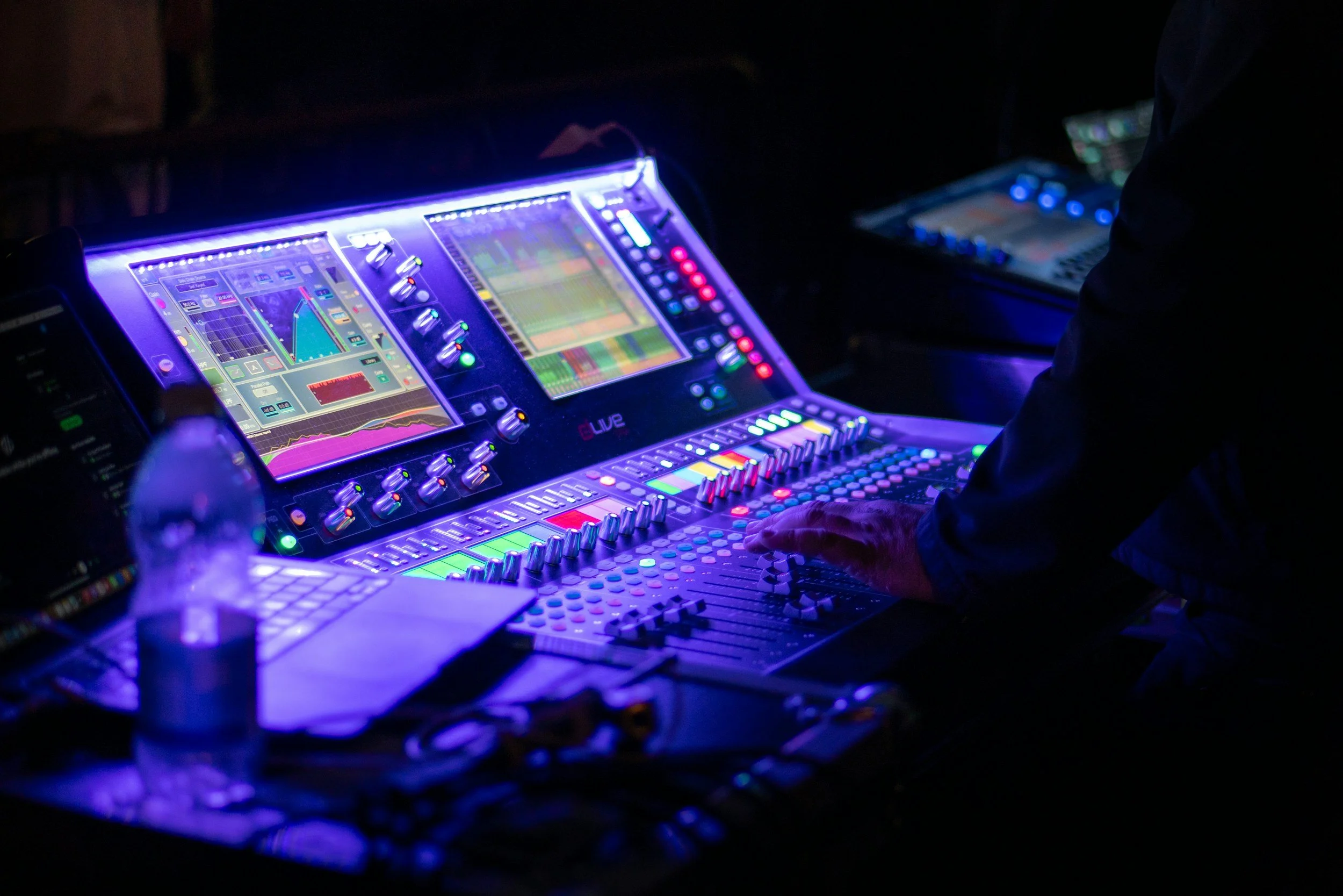 Close-up view of electronic music production equipment with colorful illuminated screens and controls at night. A person is operating the equipment, which includes a mixing console and digital interfaces, in a dark environment.