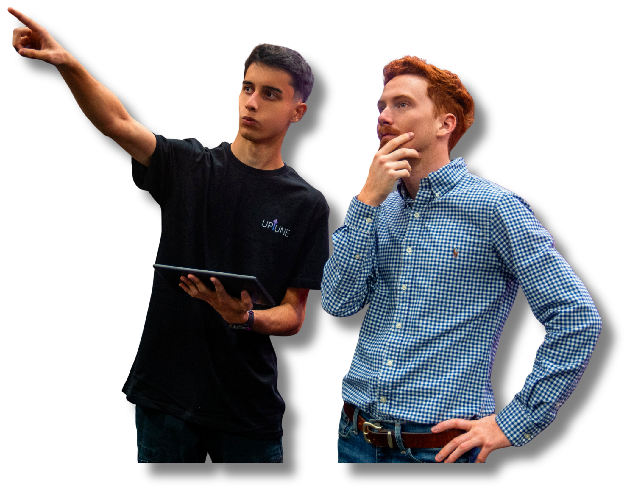 Two men are engaged in a discussion; one is pointing to the left while holding a tablet, and the other is thinking with his hand on his chin. The man on the left has dark hair and is wearing a black t-shirt with a logo, and the man on the right has red hair and is wearing a blue checkered shirt.