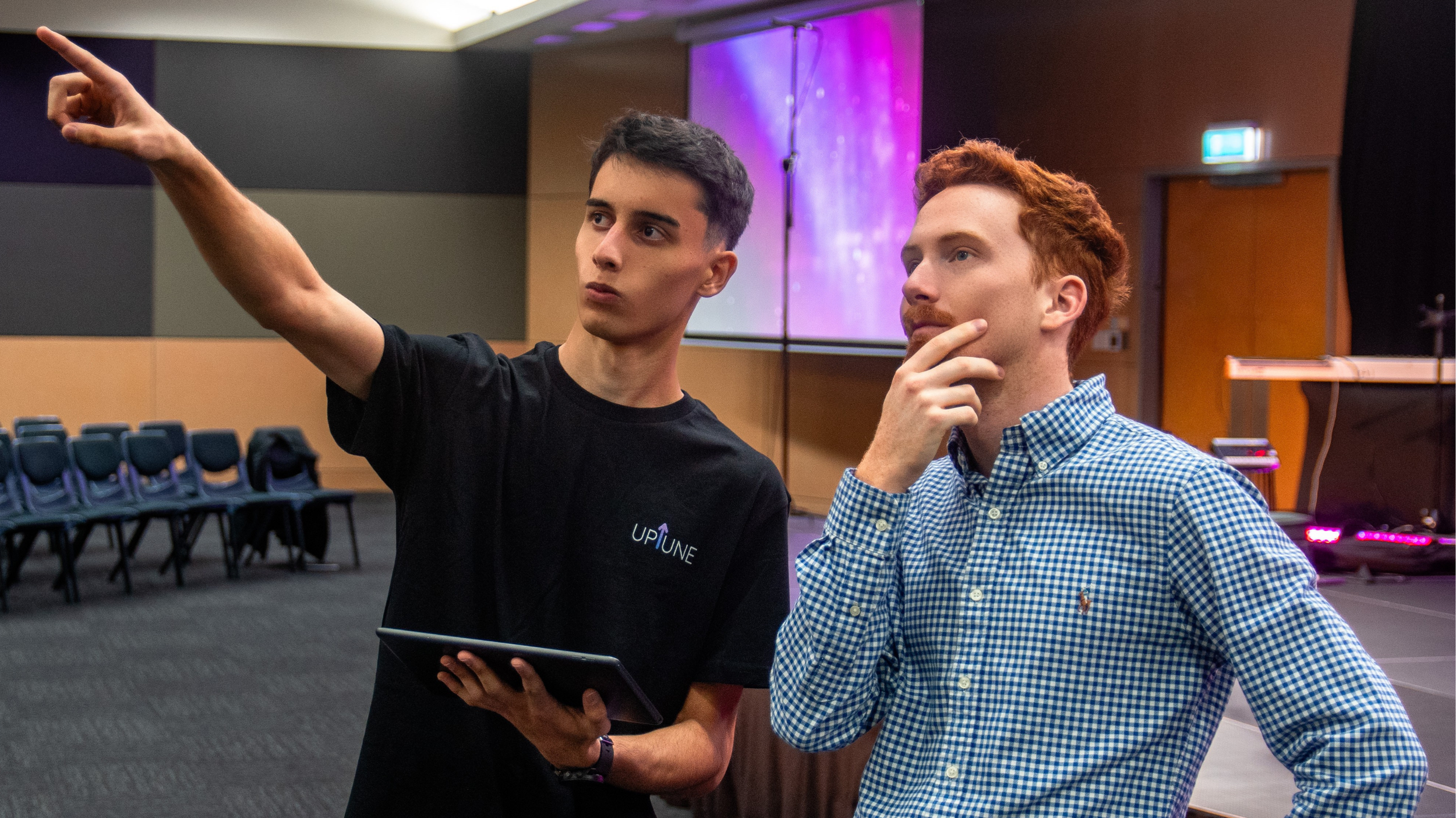 Two young men at an indoor event, one is pointing at something off-camera while the other is thoughtfully resting his chin on his hand.