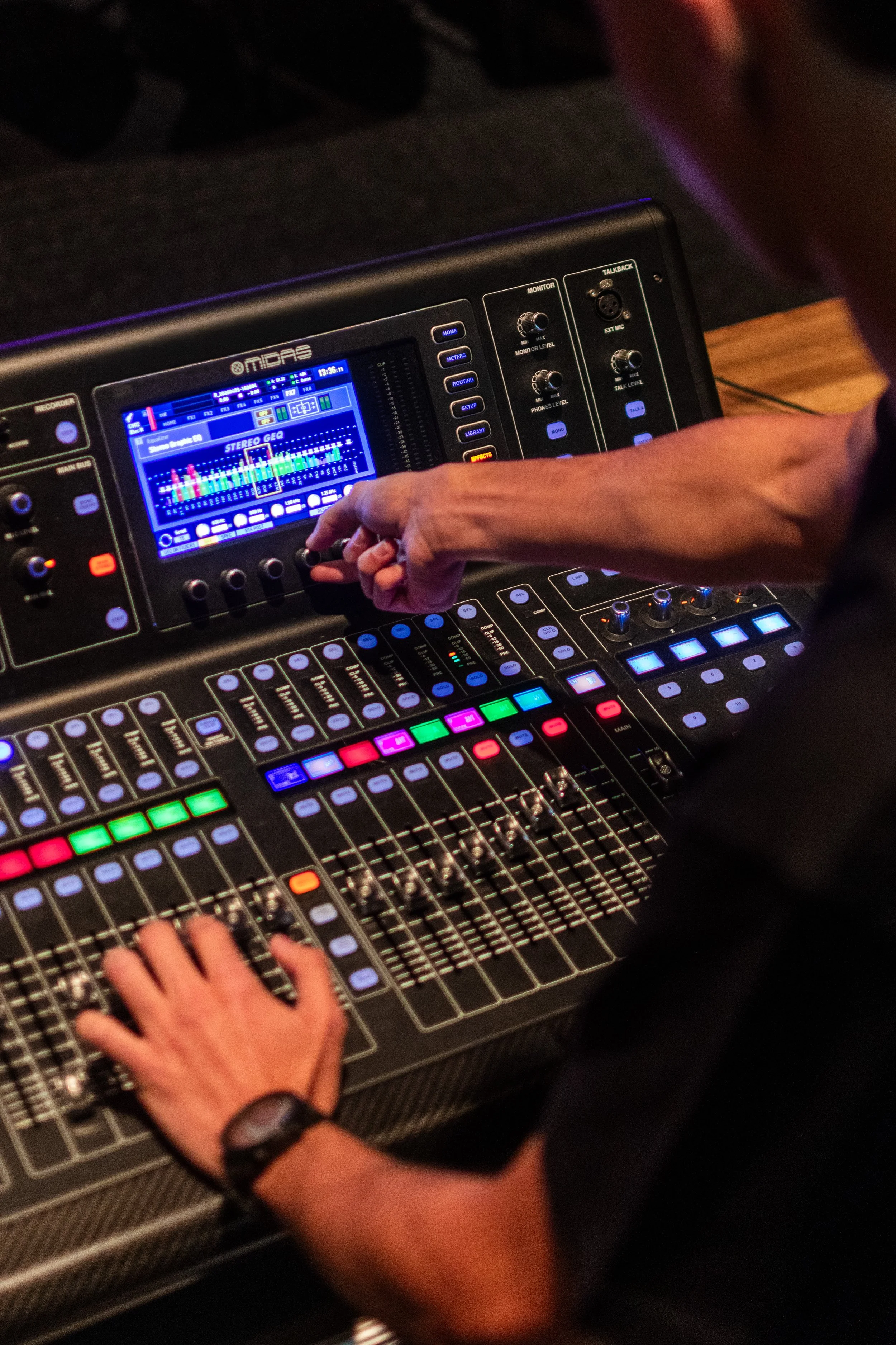 Close-up of a DJ or sound engineer adjusting a sound mixing console with various colorful buttons, sliders, and a digital display showing audio levels and settings.