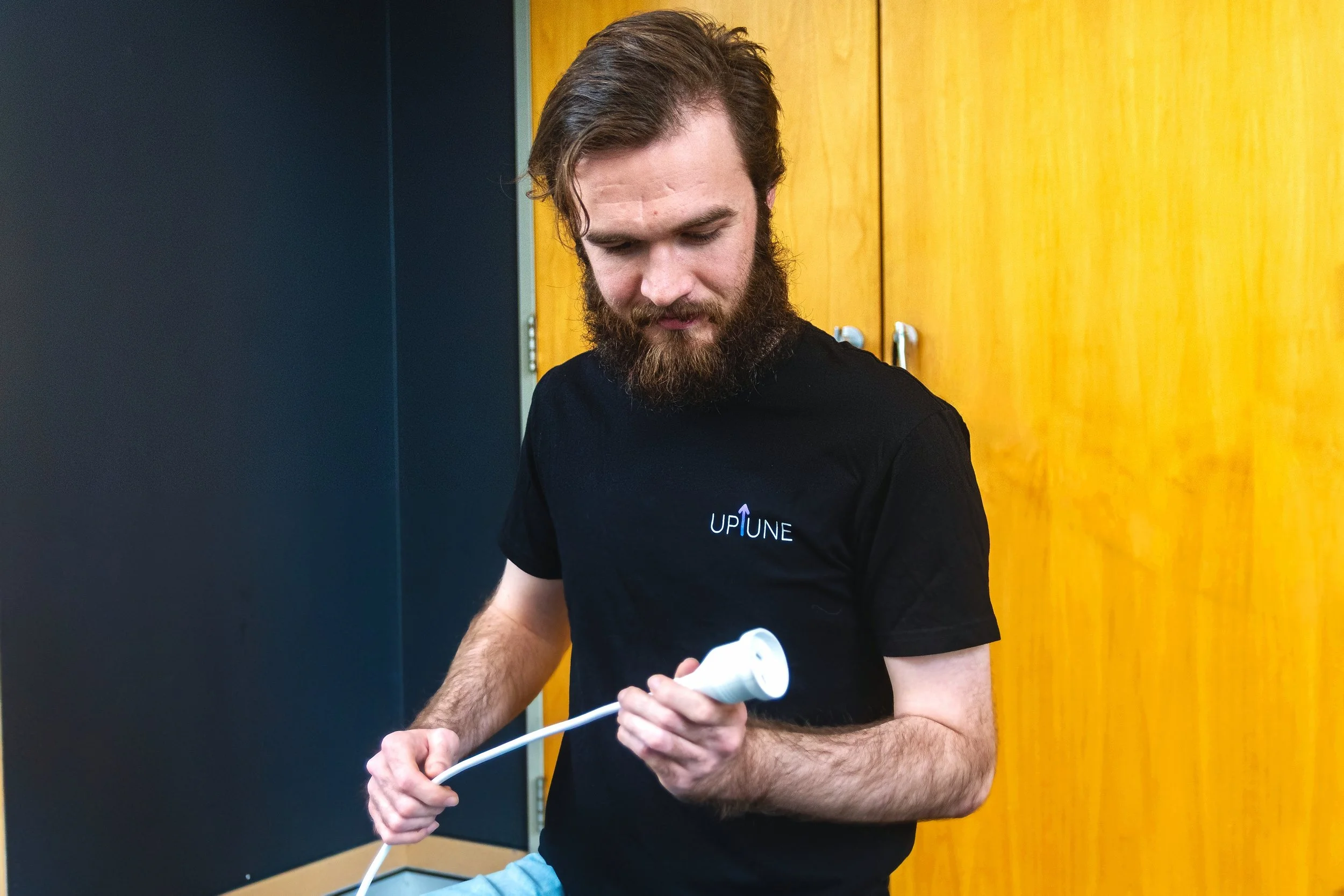 A man with a beard and long hair is holding a white cable and a small white electronic device. He is wearing a black T-shirt with the word 'UPTUNE' printed on it. The background features a yellow wooden wall and a dark-colored wall on the left.