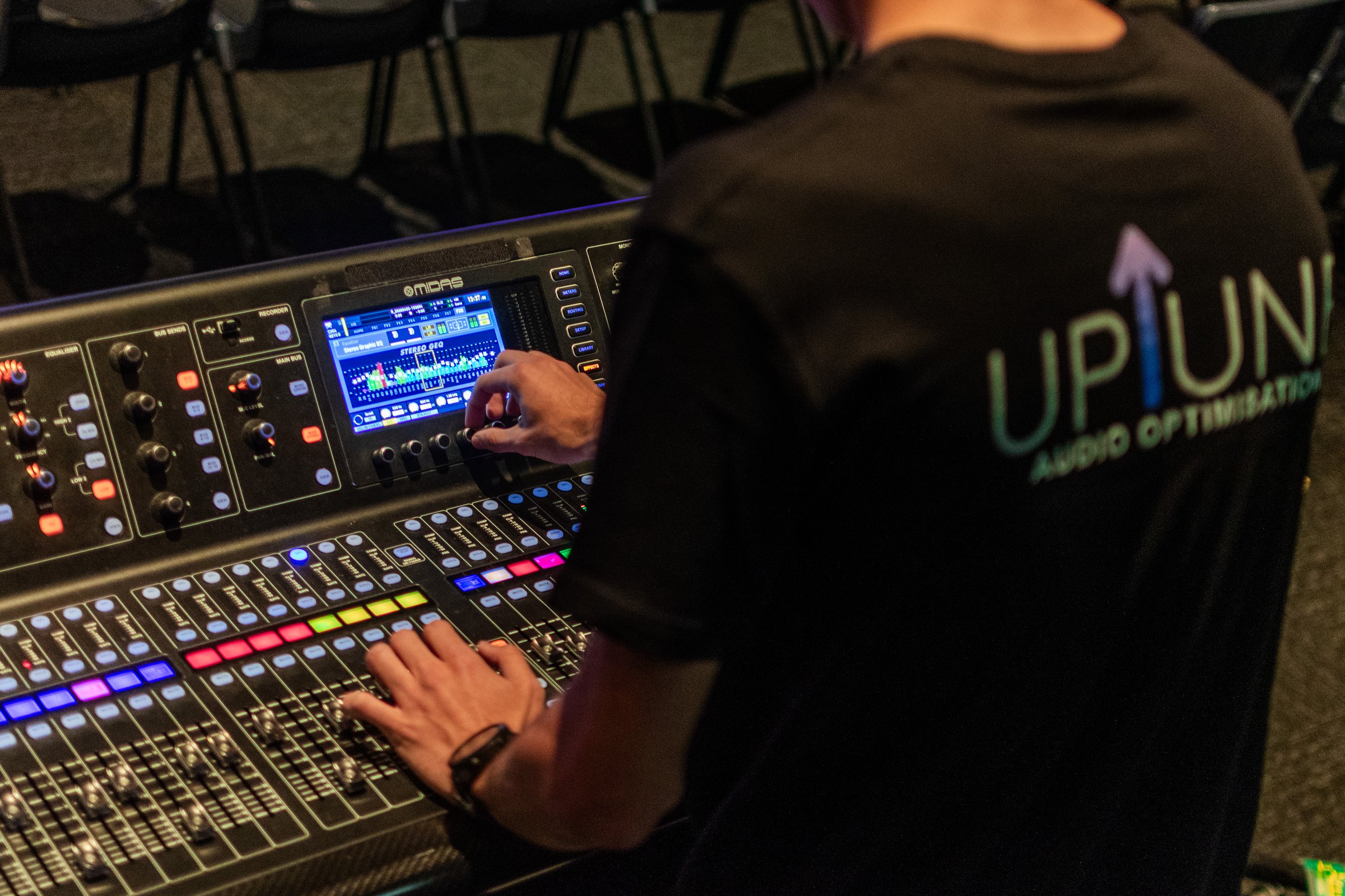 A person operating a digital sound mixing console with colorful buttons and a digital display, in a dark room with chairs in the background. The individual is wearing a shirt with the logo 'UPON AUDIO OPTIMIZATION' on the back.