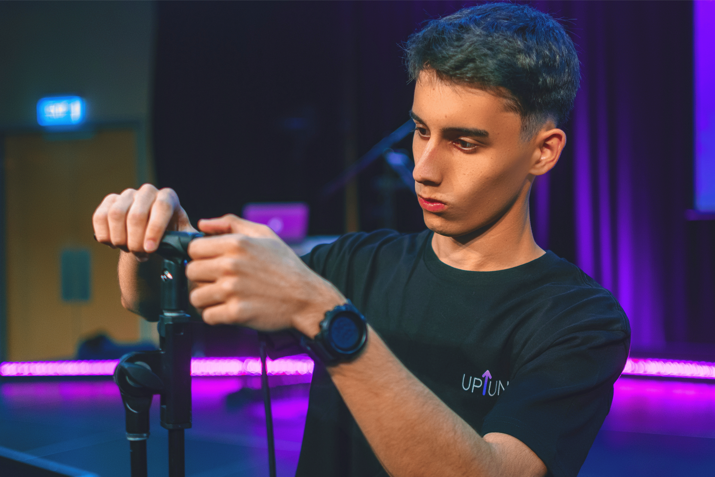 A young man with short dark hair and wearing a black T-shirt with the logo 'UP TUN' is adjusting a camera at a tripod in a room with purple and blue lighting.