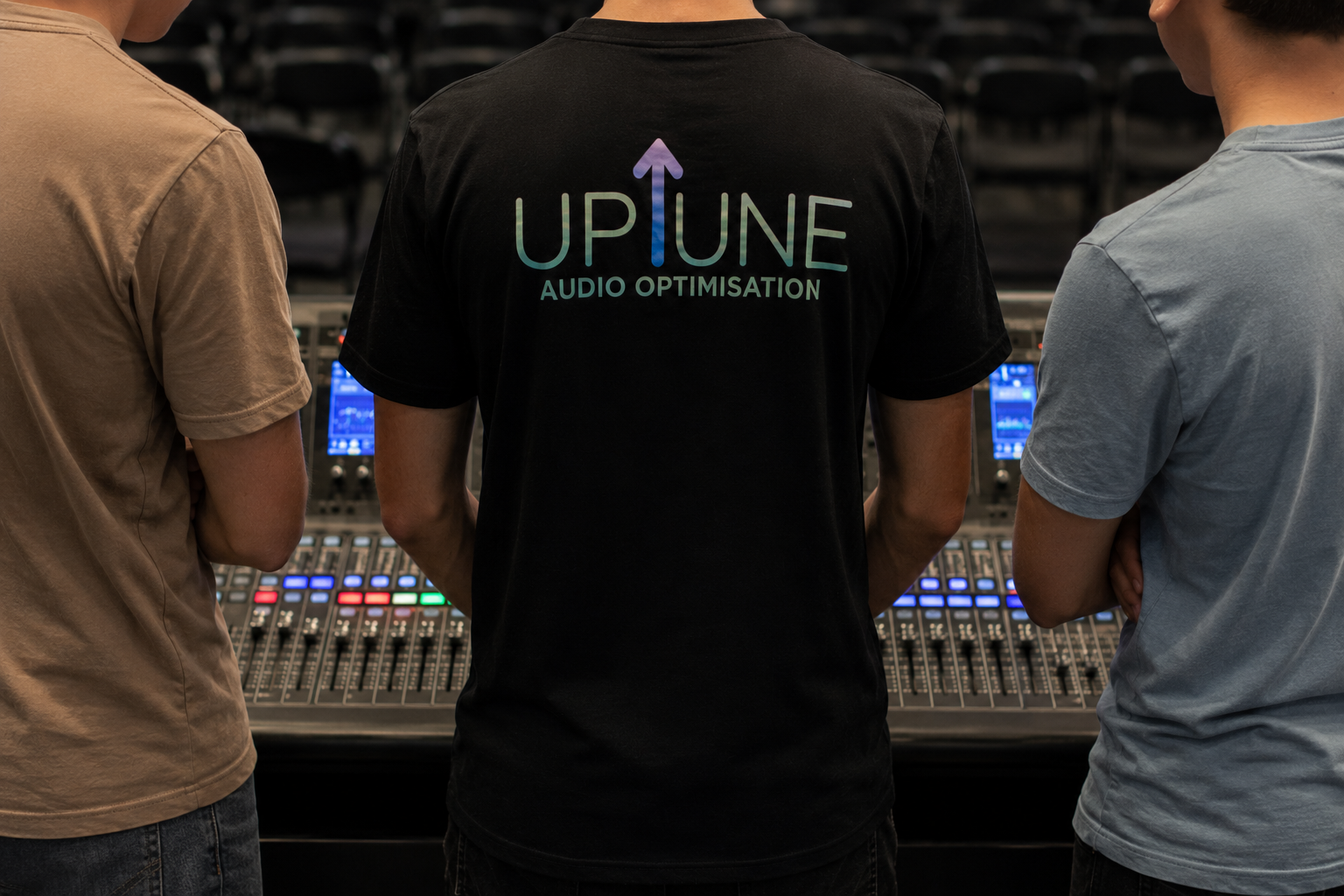 Three people standing around an audio mixing console with screens and controls, one wearing a black t-shirt with 'UPTUNE AUDIO OPTIMISATION' on the back, in a recording studio or event setting.