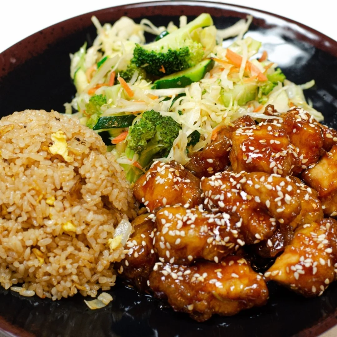Fried rice, mixed vegetables, and orange sesame chicken on a black plate.